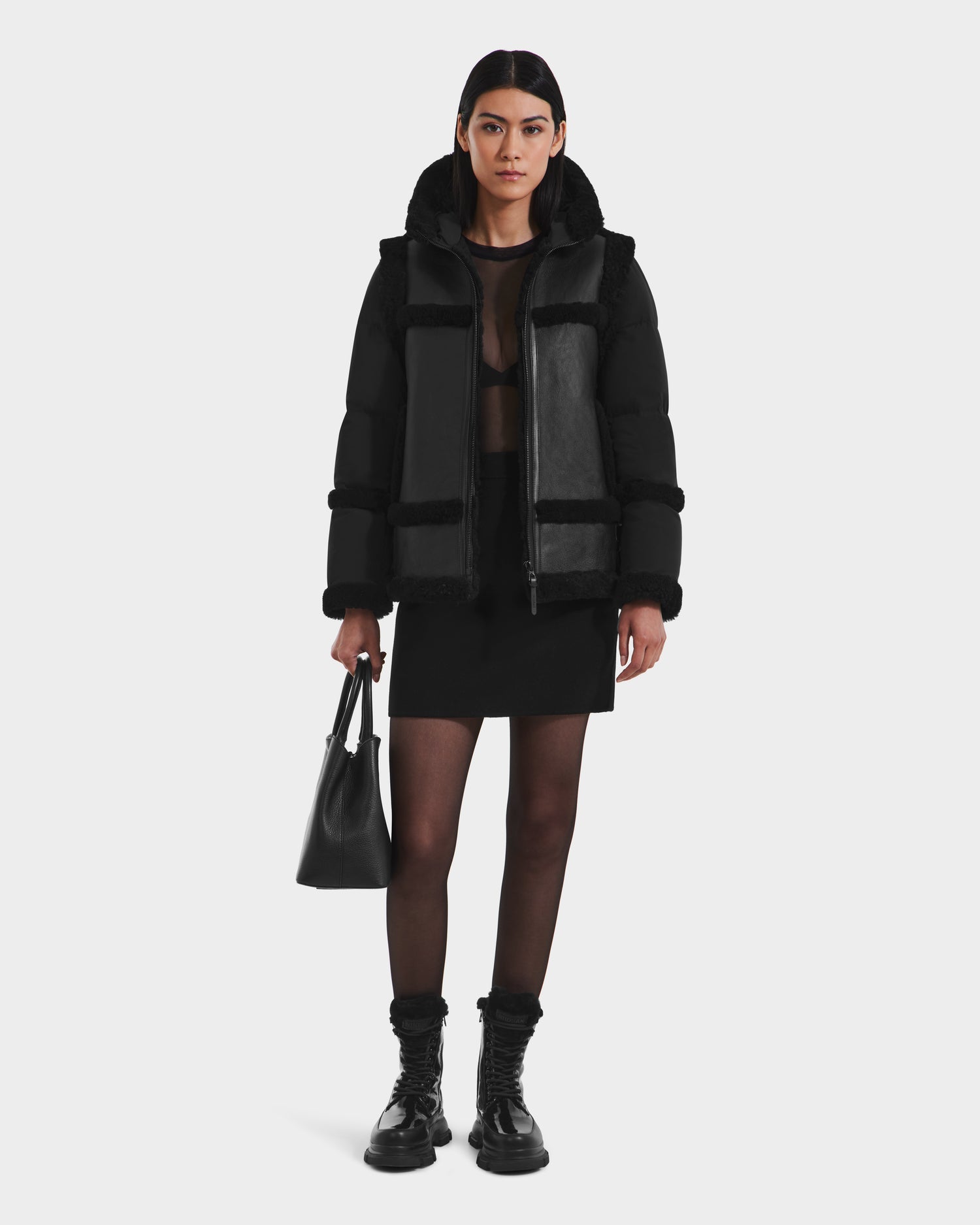 A woman with straight dark hair stands facing forward in the Rudsak PHEOBE - 8125510 Black shearling puffer, sheer top, mini skirt, tights, and combat boots. She holds a black handbag against a plain white background that highlights her outerwear.