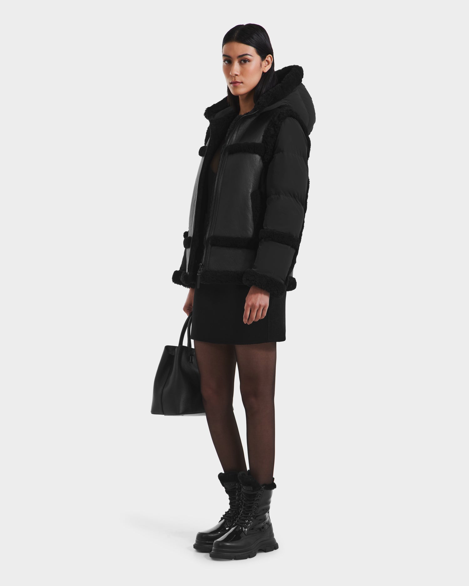 A woman faces slightly to the side, wearing the Rudsak PHEOBE - 8125510 Black puffer jacket with a hood, styled with a black mini dress, sheer black tights, chunky boots, and a black handbag against a plain white background.