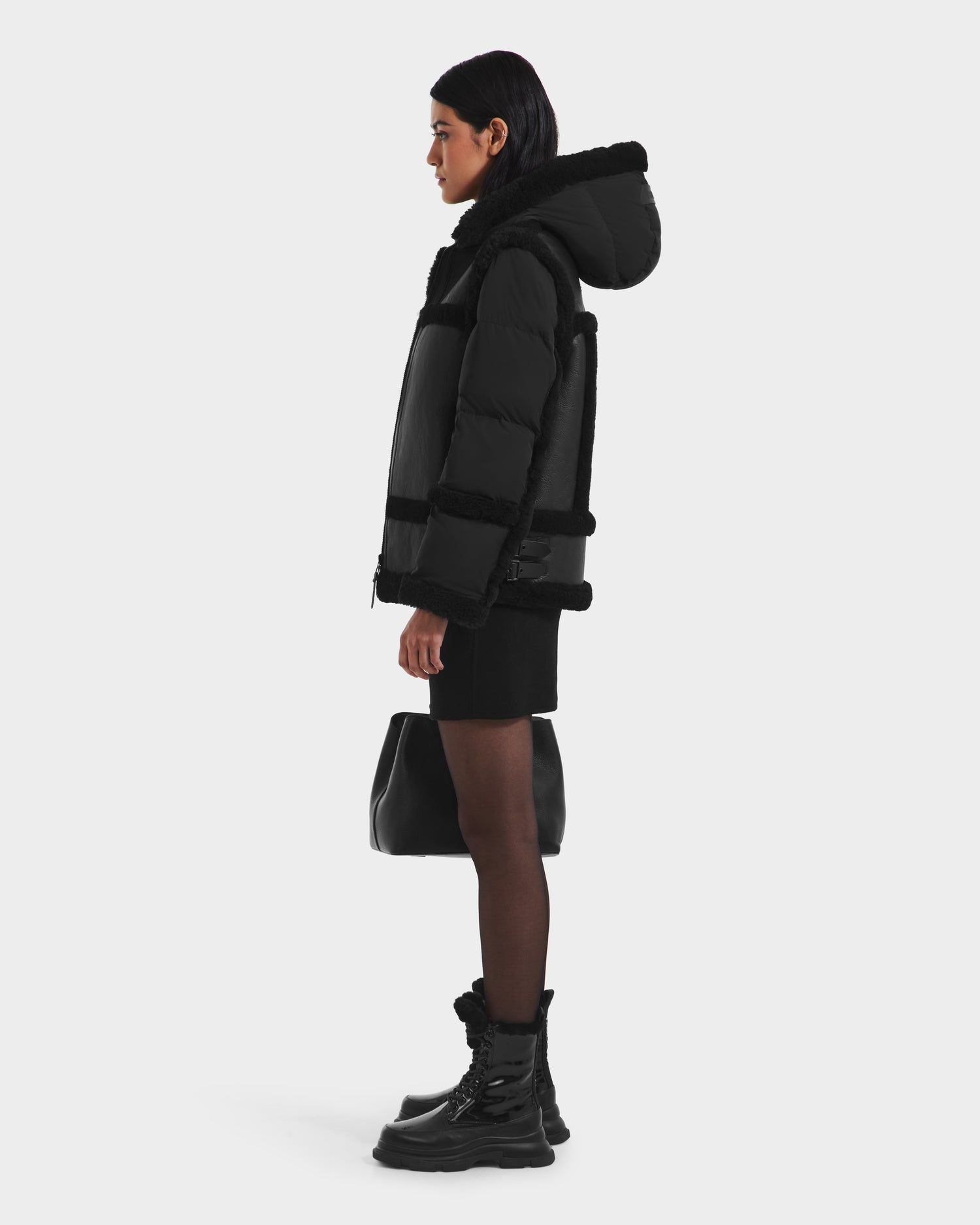 A woman in profile wears the Rudsak PHEOBE - 8125510 Black shearling puffer jacket with hood, paired with a short black skirt, tights, boots, and a black handbag—stylish transitional outerwear against a white background.