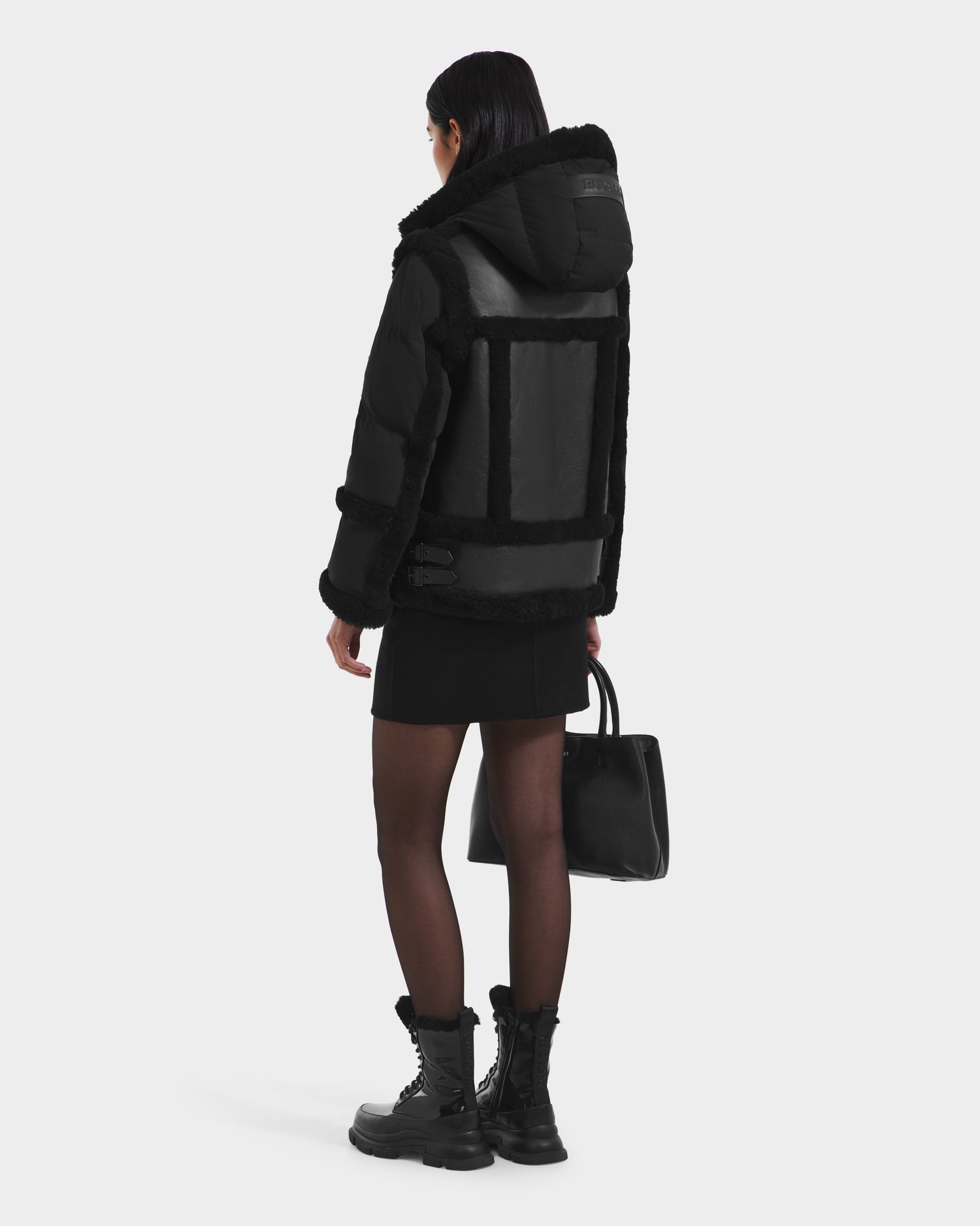 A person with long dark hair, seen from behind, wears the Rudsak PHEOBE - 8125510 Black women’s jacket with a black skirt, sheer tights, combat boots, and carries a black handbag against a plain light background—ideal transitional outerwear.