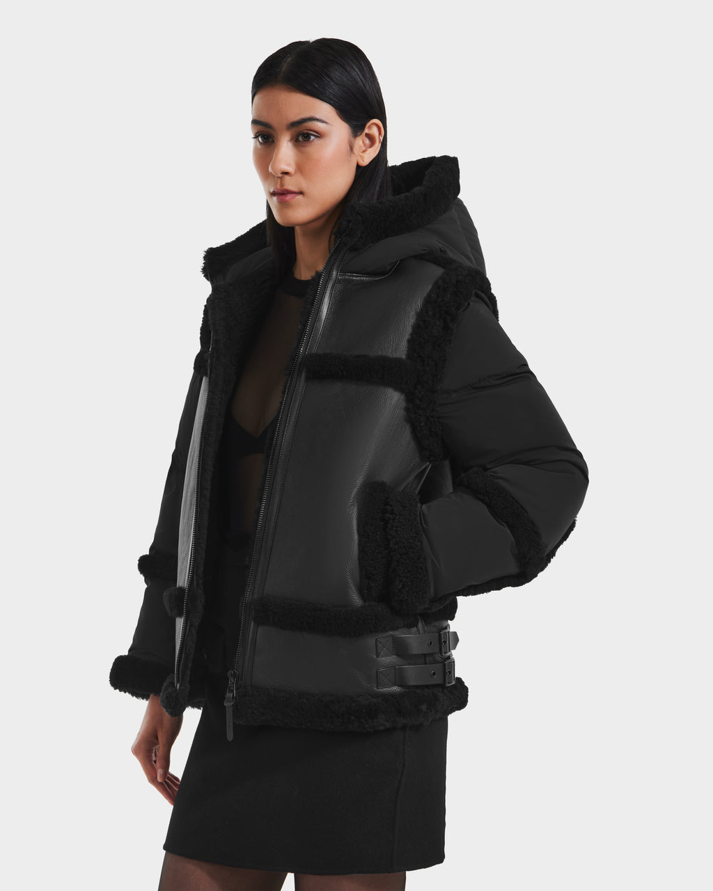 A woman wears the Rudsak PHEOBE - 8125510 Black shearling jacket with buckle details over a sheer black top and mini skirt, standing against a white background—ideal transitional style for the season.