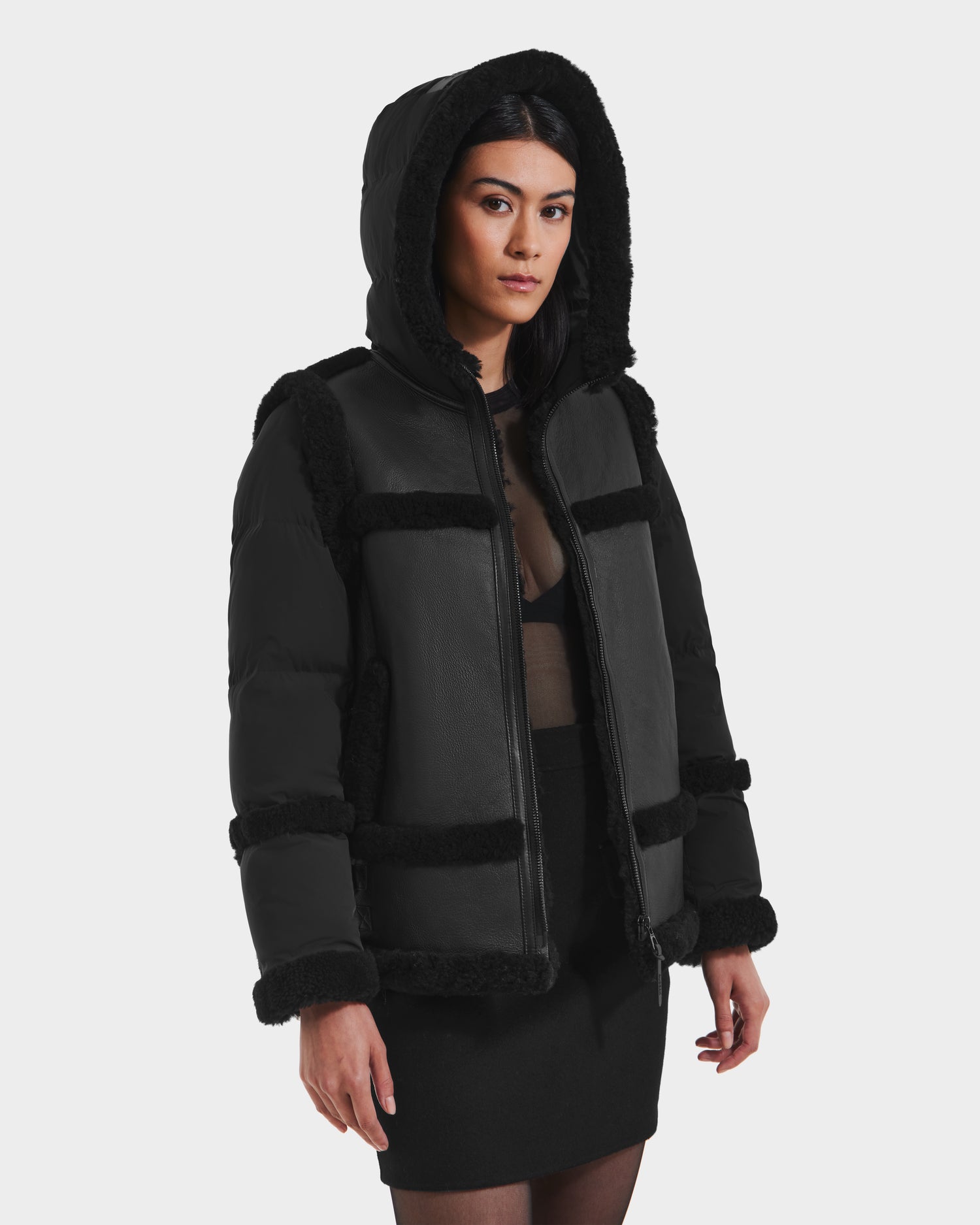 Wearing the PHEOBE - 8125510 Black jacket by Rudsak, a woman with dark hair layers this faux fur-trimmed outerwear over a sheer black top and skirt, creating a chic winter look against a white background.