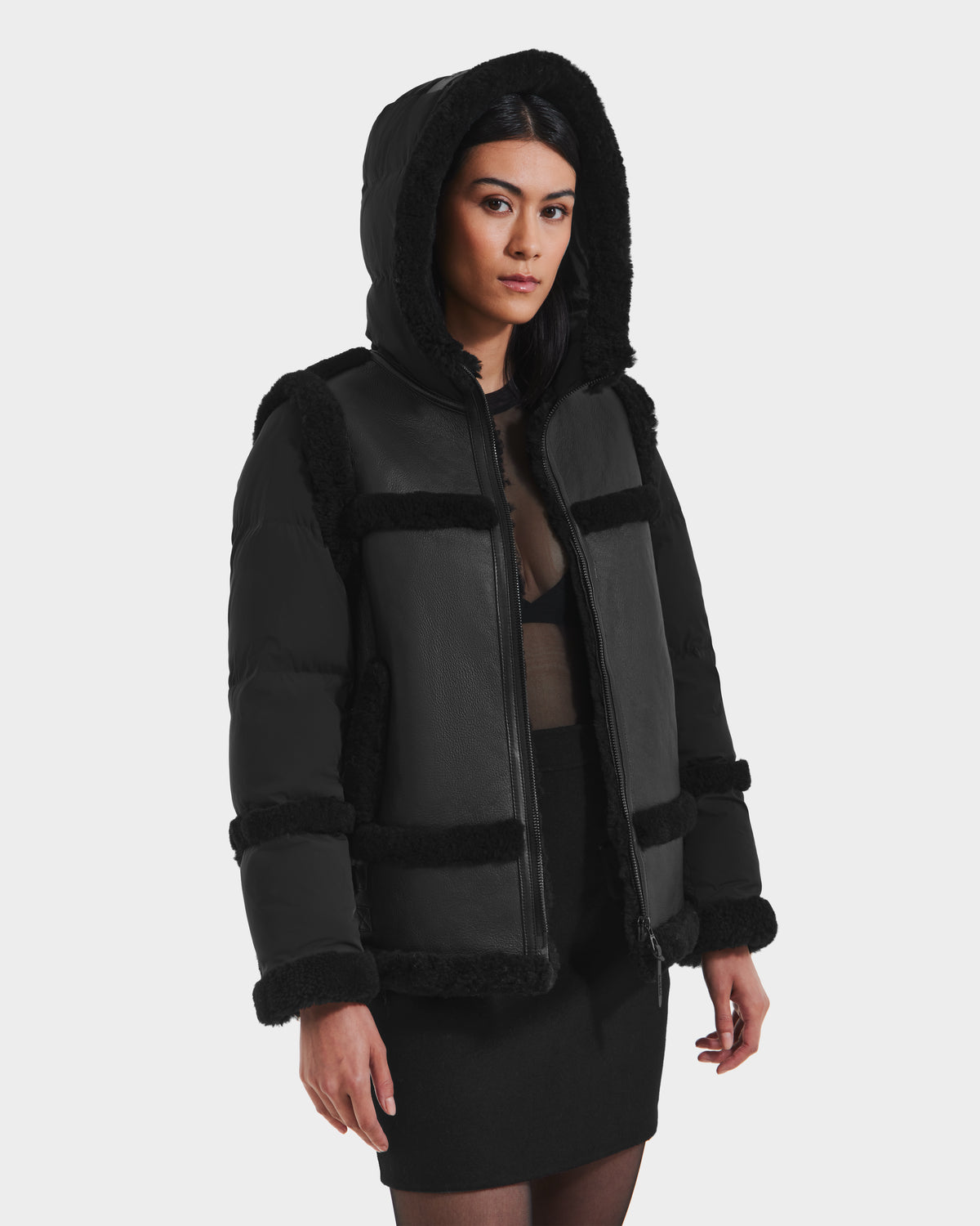 Wearing the PHEOBE - 8125510 Black jacket by Rudsak, a woman with dark hair layers this faux fur-trimmed outerwear over a sheer black top and skirt, creating a chic winter look against a white background.