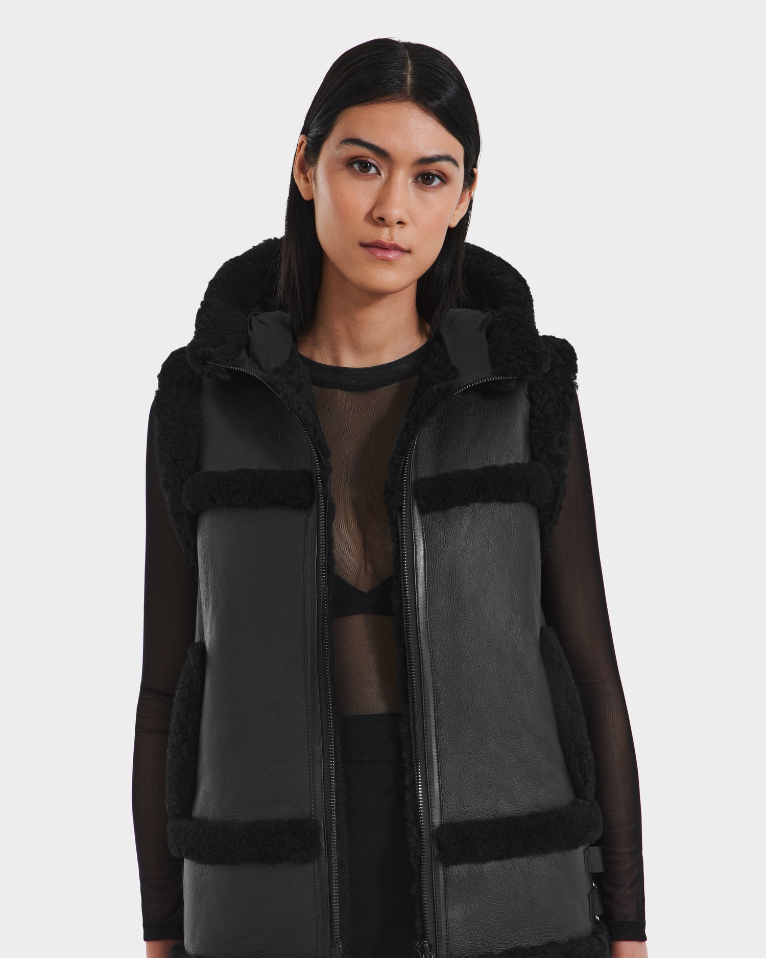 A woman with straight dark hair wears the Rudsak PHEOBE - 8125510 Black sleeveless shearling vest over a sheer black long-sleeve top, standing against a plain light background and looking directly at the camera.