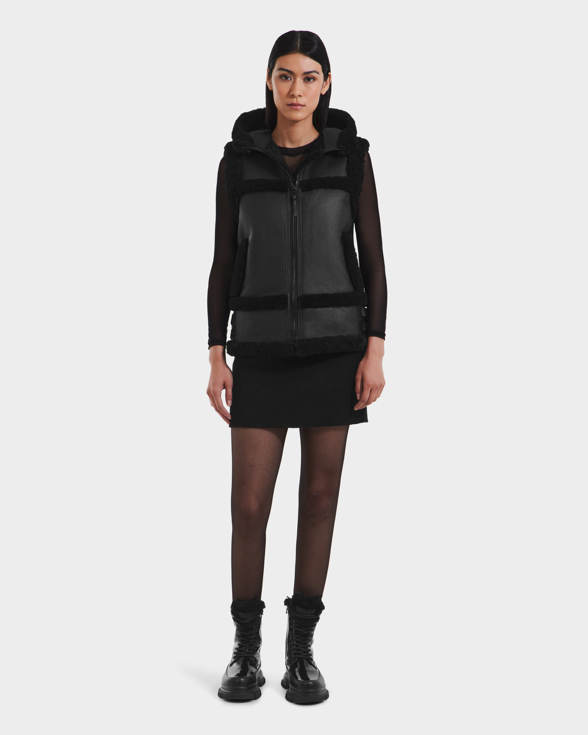 A woman faces forward wearing the PHEOBE - 8125510 Black vest by Rudsak, styled with a black skirt, sheer black tights, and black boots against a plain white background.