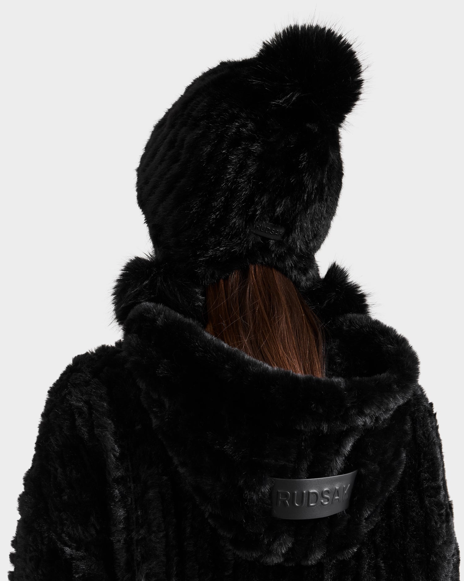 A person with long brown hair wears the Rudsak PIA - 8425547 Black faux fur hooded coat and a matching women’s faux fur beanie with pom-pom. The coat’s back has a black patch with "RUDSAK" written on it.