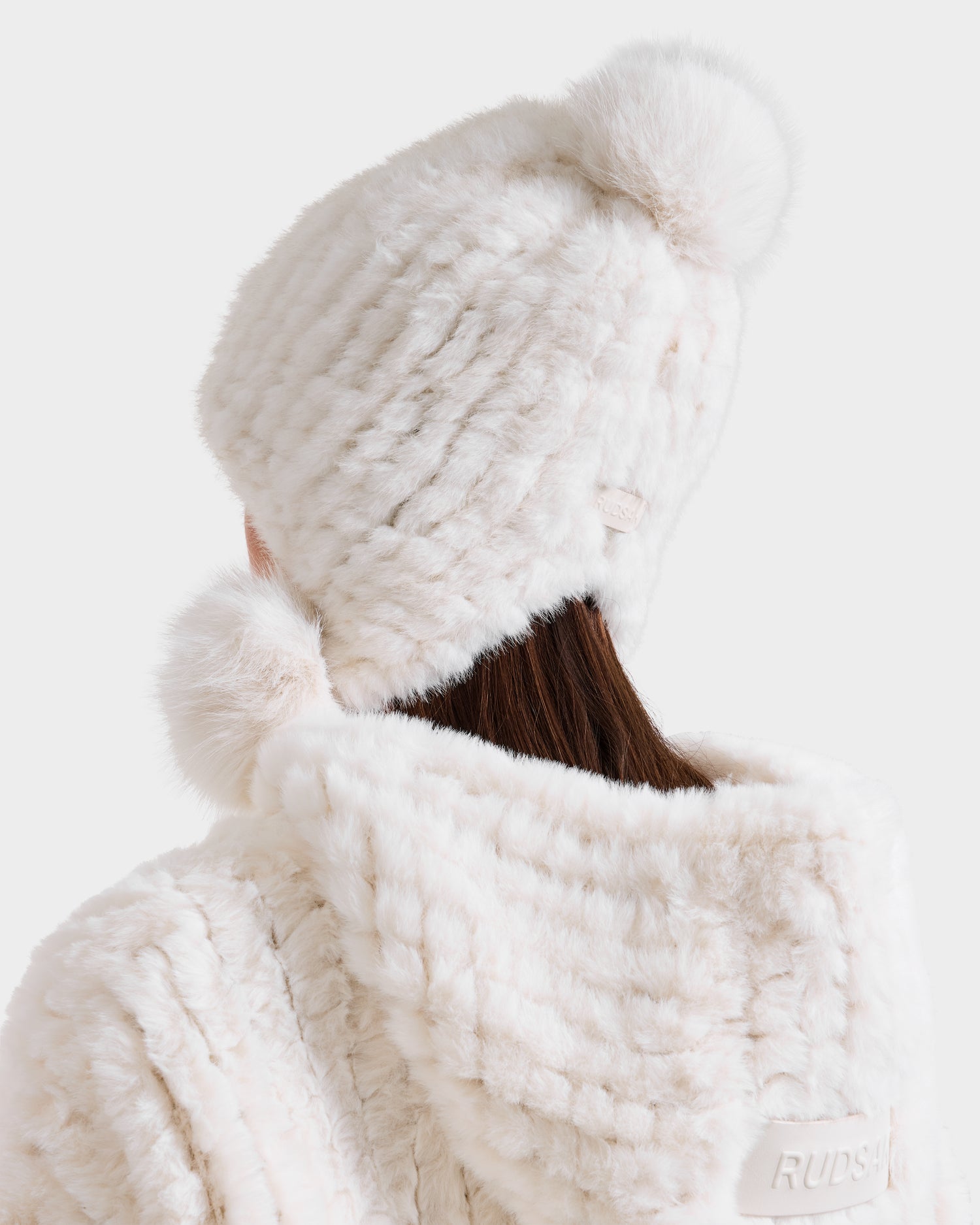 A person with long brown hair is seen from behind wearing the Rudsak PIA - 8425547 Iceland women’s faux fur hat with ear flaps and pom-pom, paired with a matching white Rudsak faux fur coat, against a white background.