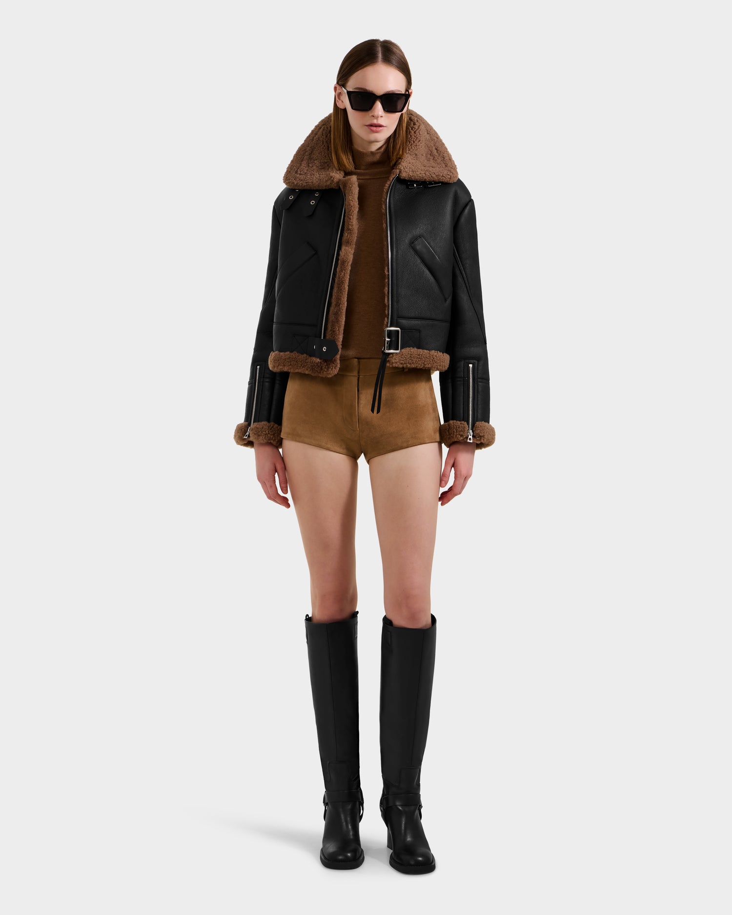 A woman in dark sunglasses and the Rudsak POLINA - 8124811 Black women's shearling jacket with brown fur trim, paired with a brown turtleneck, brown shorts, and knee-high black boots stands against a plain light background.
