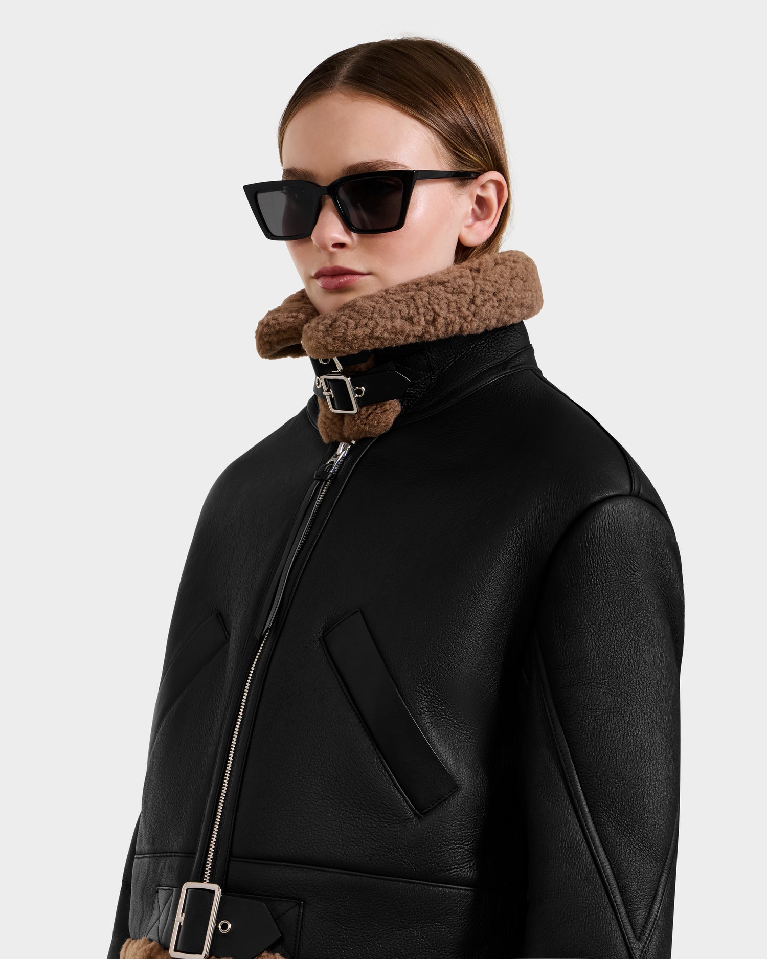 A person wearing Rudsak's POLINA - 8124811 Black oversized sunglasses and a women's shearling moto jacket with belted details, standing against a plain light background.