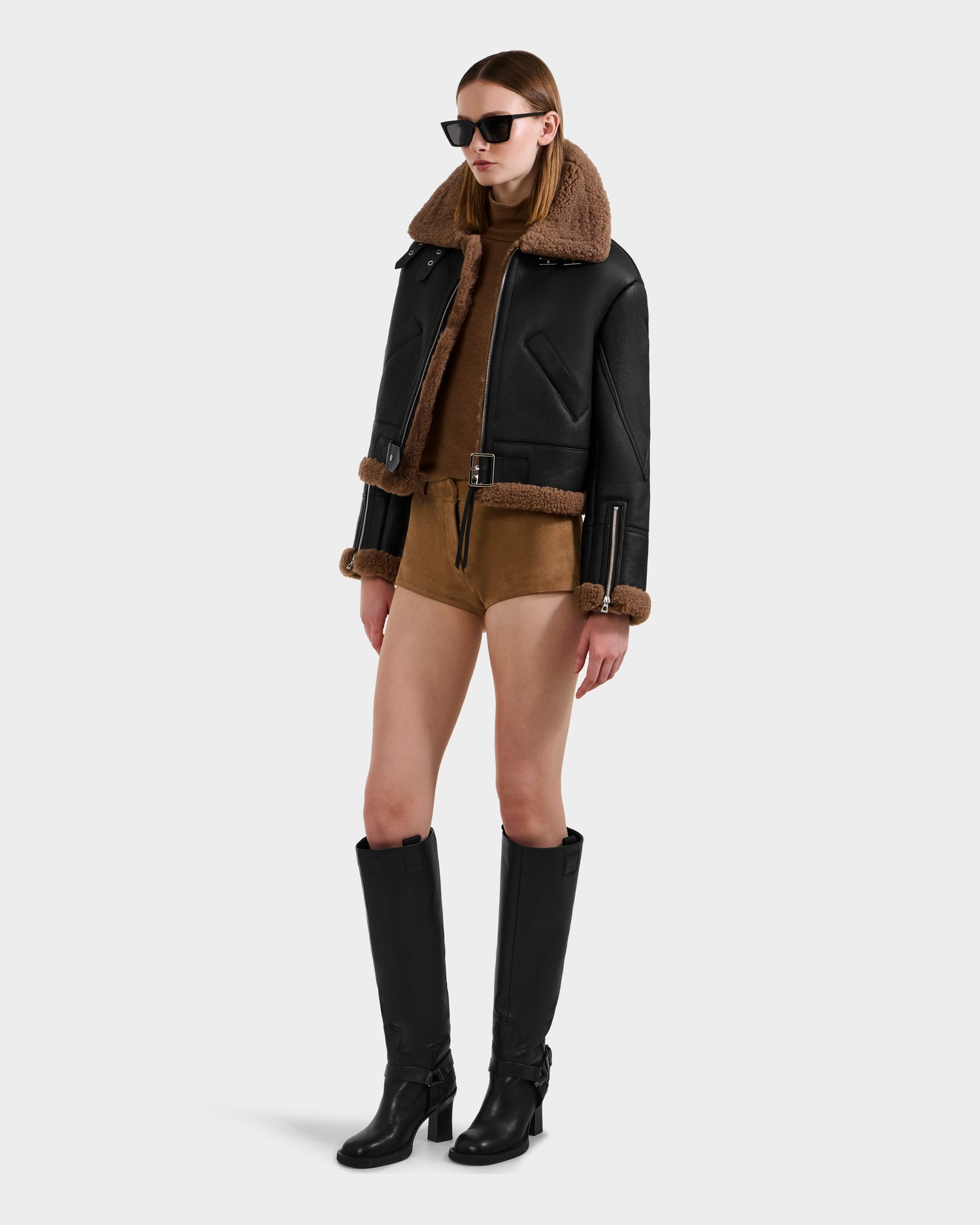 A woman wearing Rudsak's POLINA - 8124811 Black leather moto jacket with brown shearling trim, a brown romper, sunglasses, and black knee-high boots stands against a plain white background.