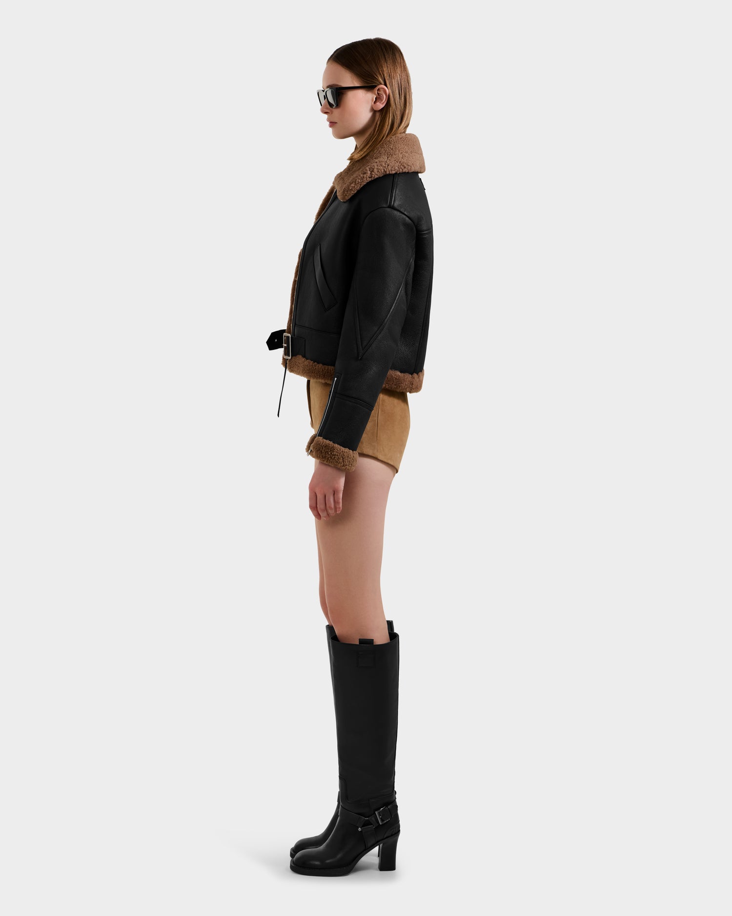 A woman in profile wears sunglasses, the Rudsak POLINA - 8124811 Black women's shearling jacket, tan shorts, and tall black heeled boots against a plain light background.