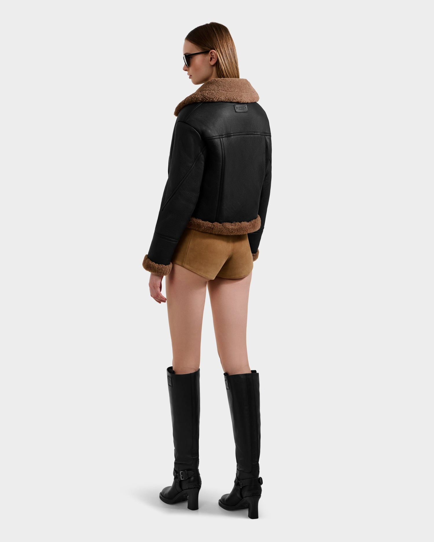 A woman faces away, wearing the Rudsak POLINA - 8124811 Black women's shearling jacket with brown shorts, knee-high black boots, and sunglasses against a plain light background.