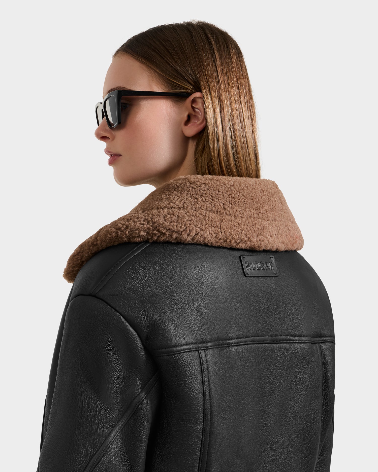 A woman with straight light brown hair, wearing black sunglasses and the Rudsak POLINA - 8124811 Black leather moto jacket with a tan shearling collar, faces away from the camera against a plain light background.