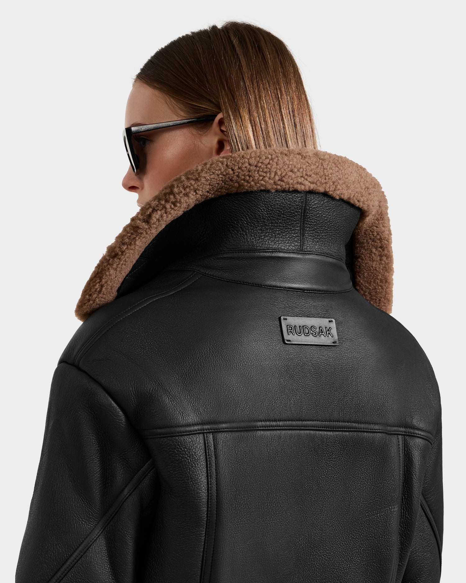A person with straight, light brown hair wears sunglasses and the Rudsak POLINA - 8124811 Black leather moto jacket with a brown shearling collar. The back of this short jacket features a rectangular “RUCAR” patch.