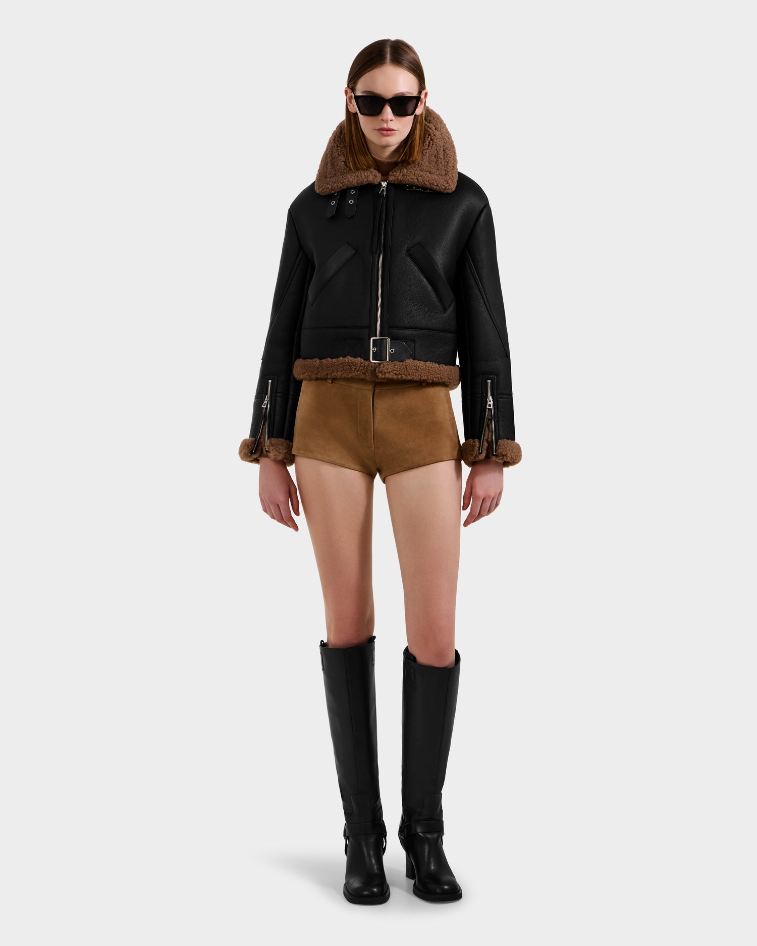 A woman in sunglasses, a black Rudsak POLINA - 8124811 shearling jacket, brown high-waisted shorts, and knee-high black boots stands against a plain white background.