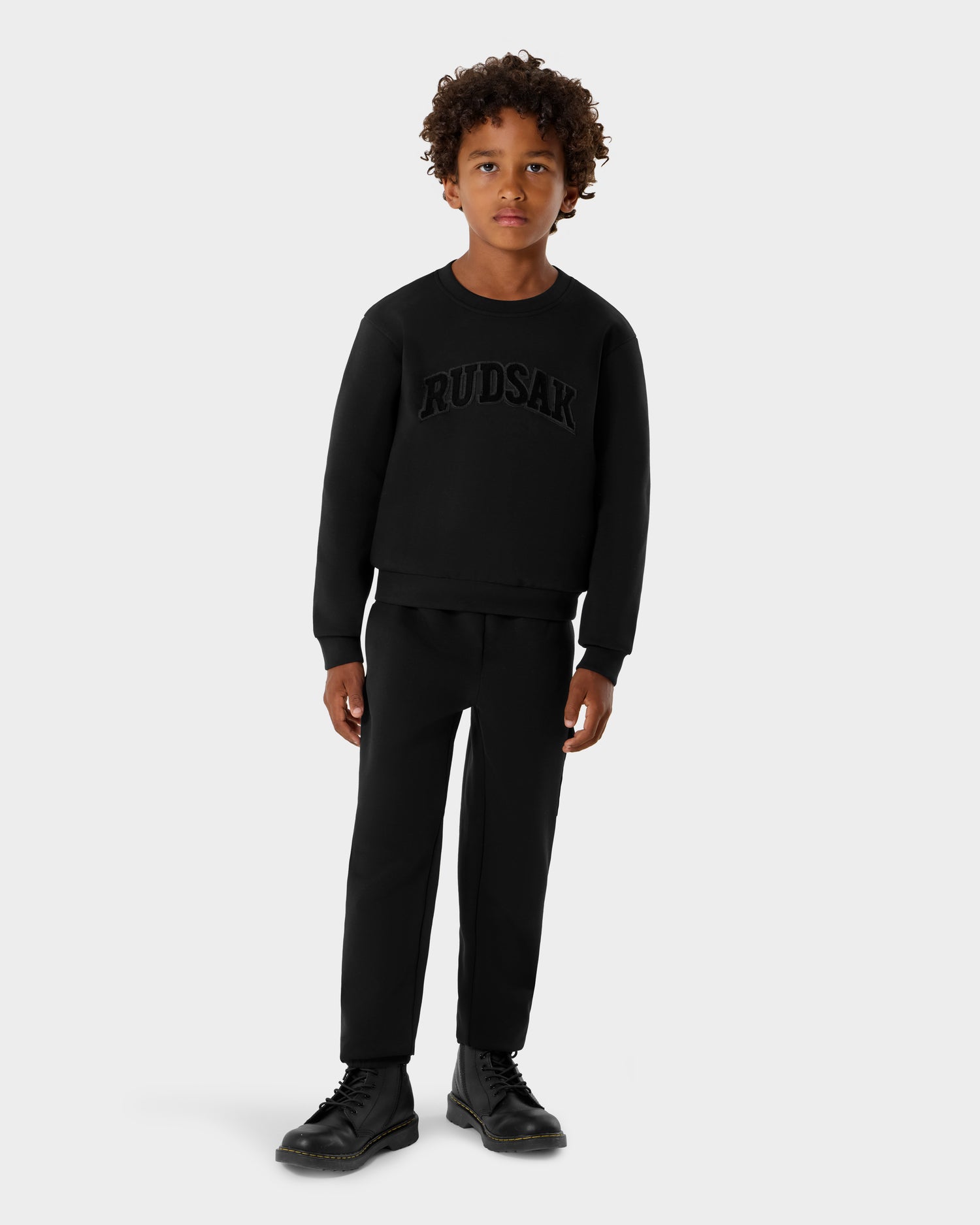 A young boy with curly hair stands facing forward in a black Rudsak POPPY - 1525003 crewneck sweatshirt, matching pants, and black boots against a plain light gray background.