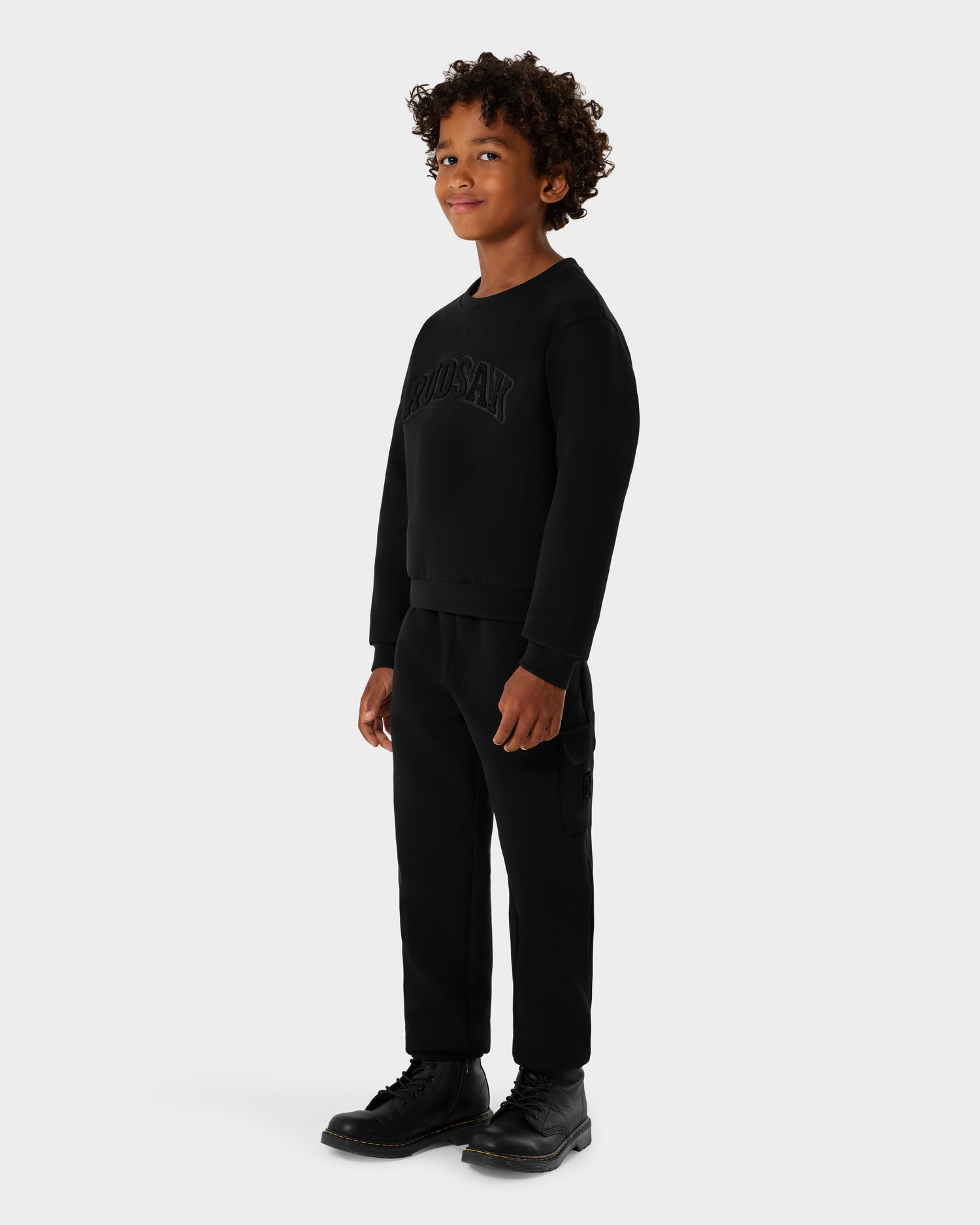 A young boy with curly hair stands against a plain background, wearing the Rudsak POPPY - 1525003 Black crewneck cotton jersey sweatshirt, paired with black pants and black boots. He faces slightly to the side and smiles softly.