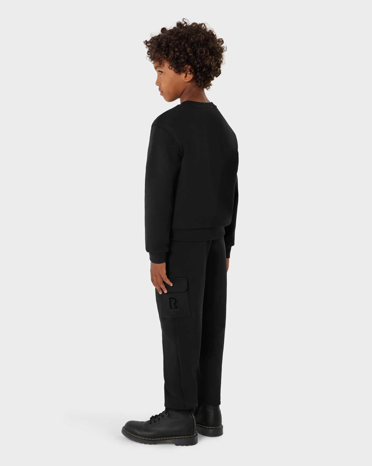 A young boy with curly hair stands at an angle, facing away, wearing the black Rudsak POPPY - 1525003 crewneck sweatshirt against a plain light gray background.
