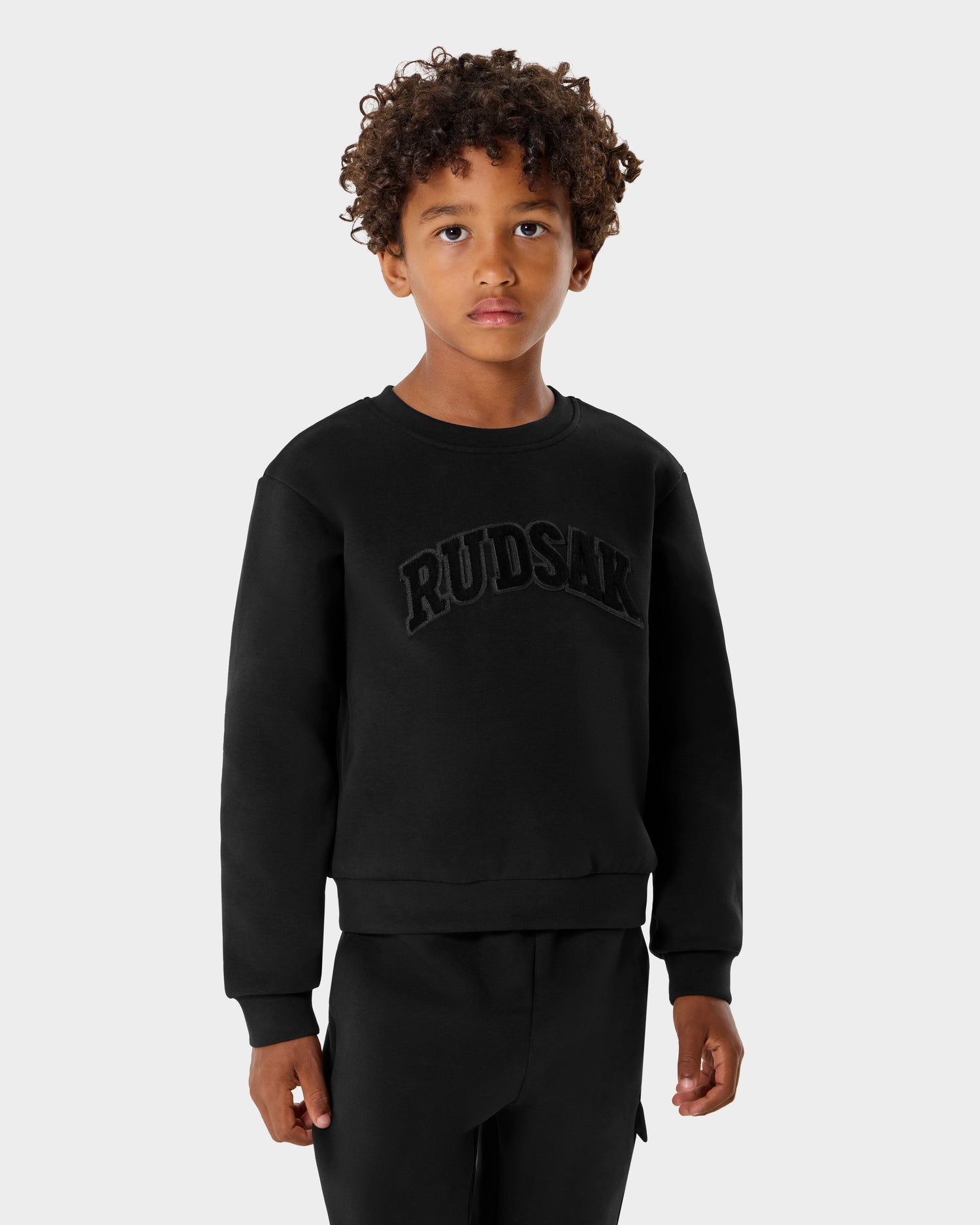 A young child with curly hair wears the Rudsak POPPY - 1525003 Black cotton crewneck sweatshirt and matching pants, featuring textured RUDSAK lettering on the front. The background is plain and light gray.