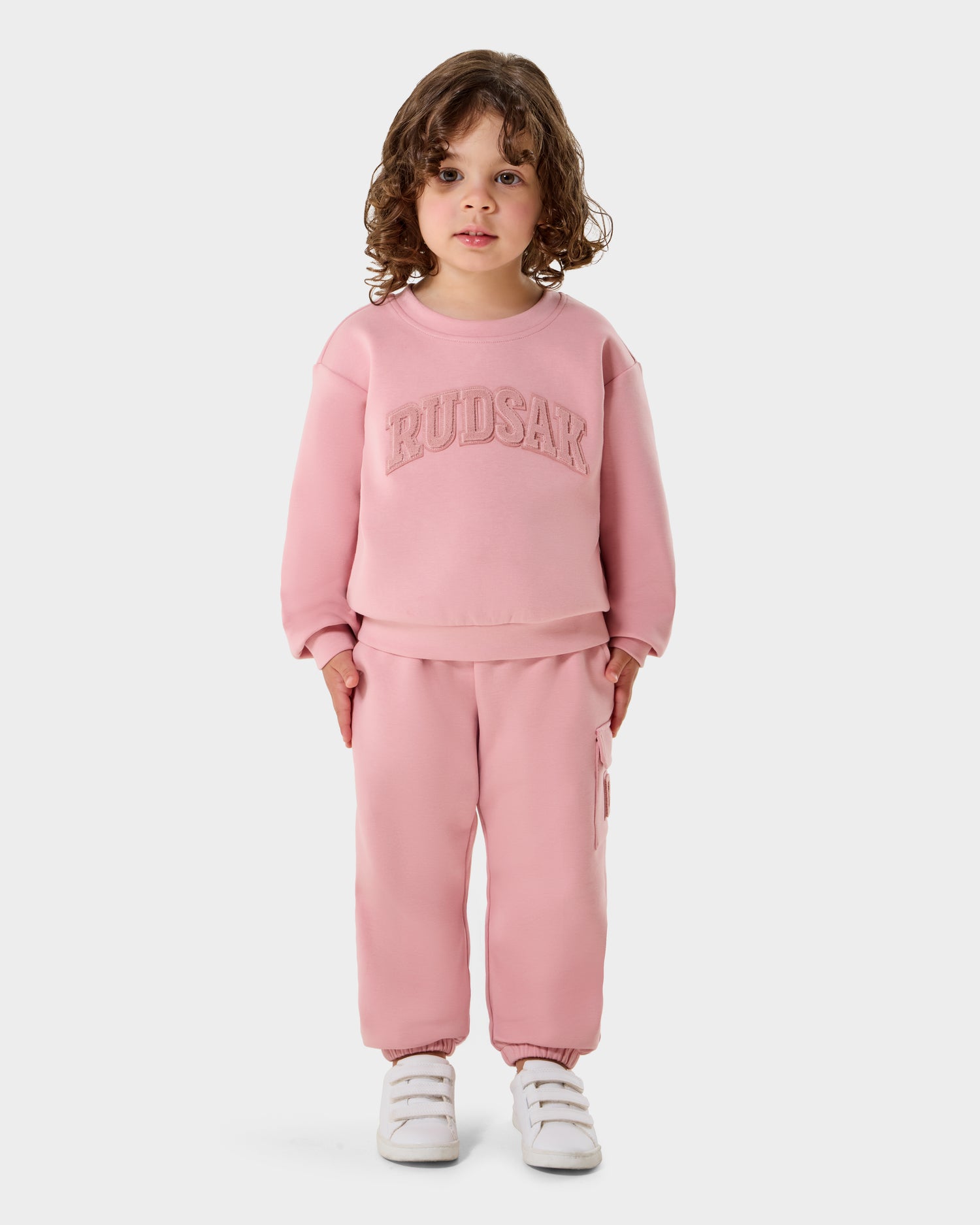 A young child with curly brown hair wears the Rudsak POPPY - 1525003 Light Pink crewneck sweatshirt and matching sweatpants, paired with white sneakers, standing against a plain white background.
