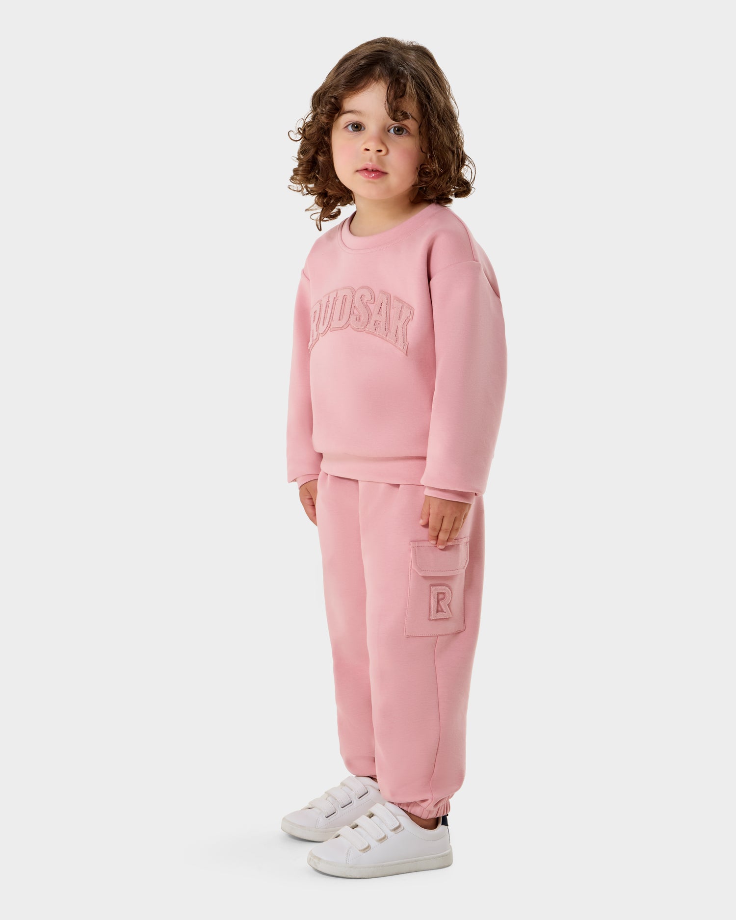 A young child with curly hair wears the Rudsak POPPY - 1525003 light pink crewneck sweatshirt and pants set with white sneakers, standing against a plain white background.