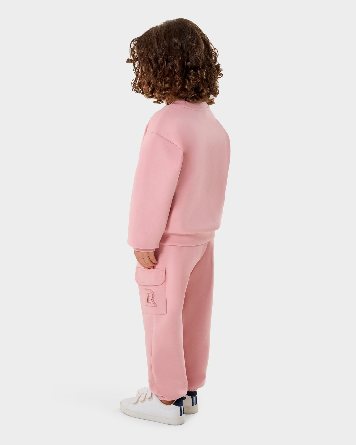 A young child with curly brown hair, facing away, wears the Rudsak POPPY - 1525003 Light Pink kids cotton sweatshirt and matching pants, paired with white sneakers with blue accents. The background is plain white.