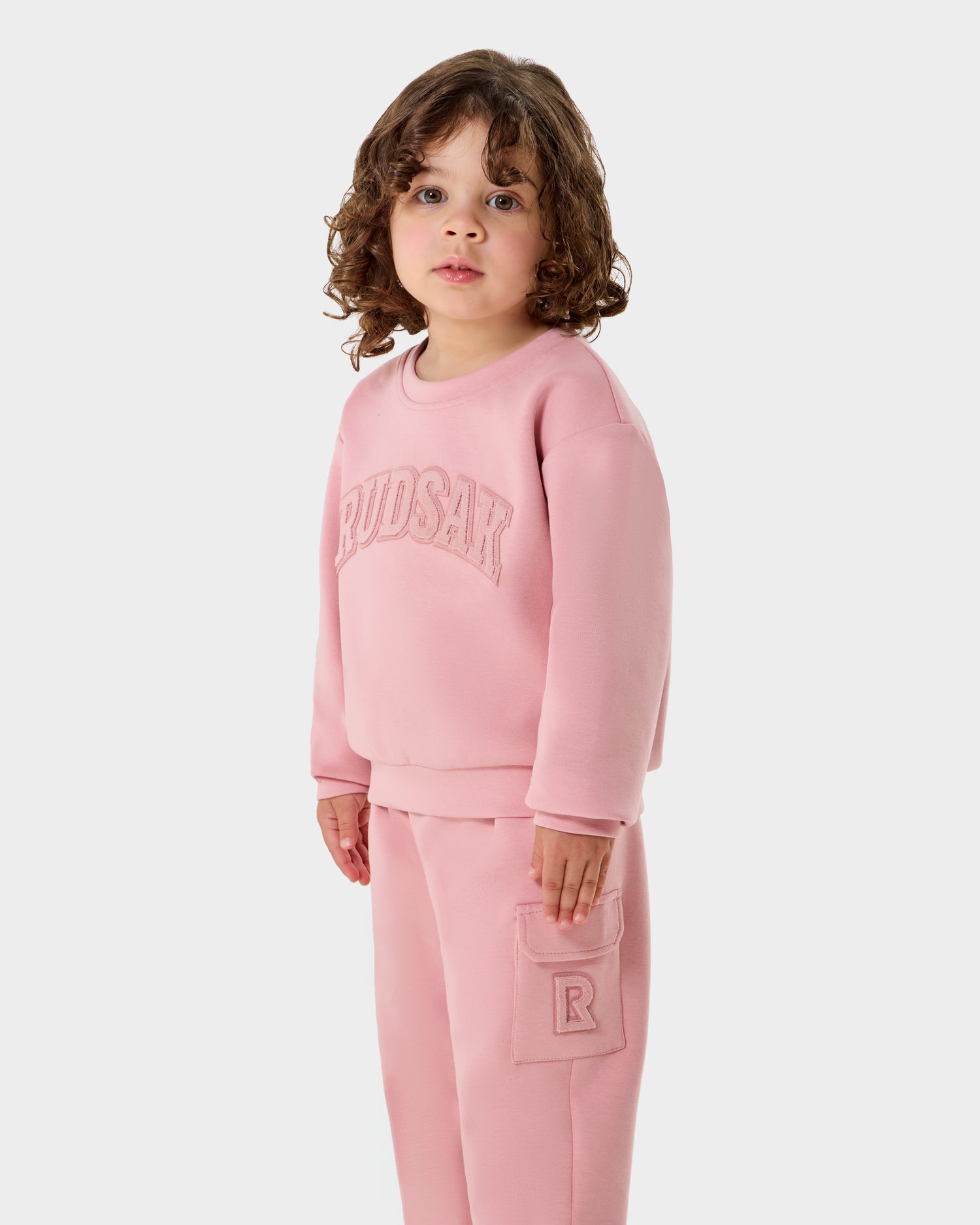 A young child with curly brown hair wears the Rudsak POPPY - 1525003 light pink kids cotton crewneck sweatshirt with "RUDSAK" on the front and matching pants with a patch pocket, standing against a plain white background.