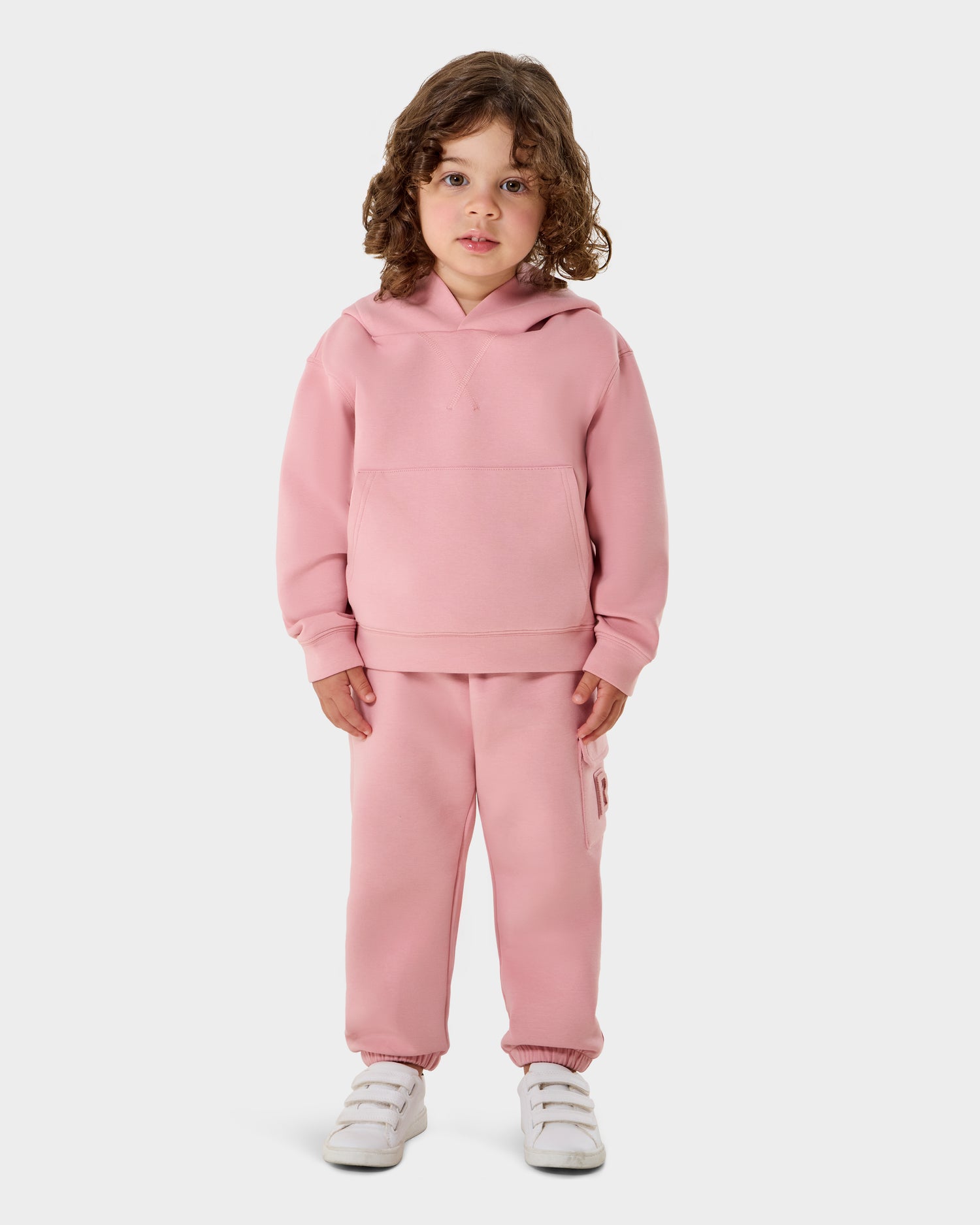 A young child with curly brown hair faces forward, wearing the Rudsak RAINEY - 1525002 hoodie and matching sweatpants in light pink, paired with white sneakers against a plain white background.