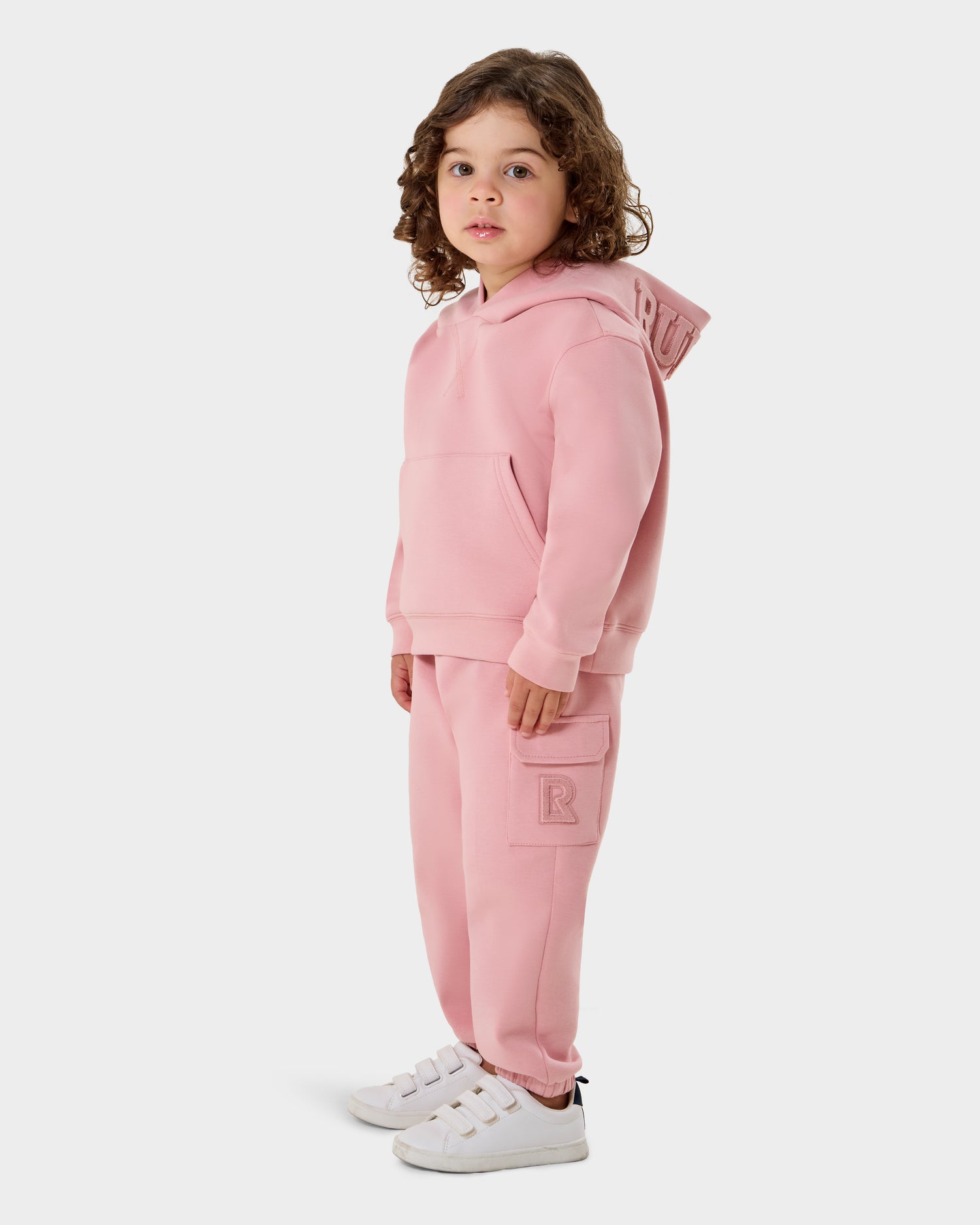 A young child with curly hair stands against a white background, wearing the Rudsak RAINEY - 1525002 light pink hoodie and sweatpants set, paired with white sneakers.