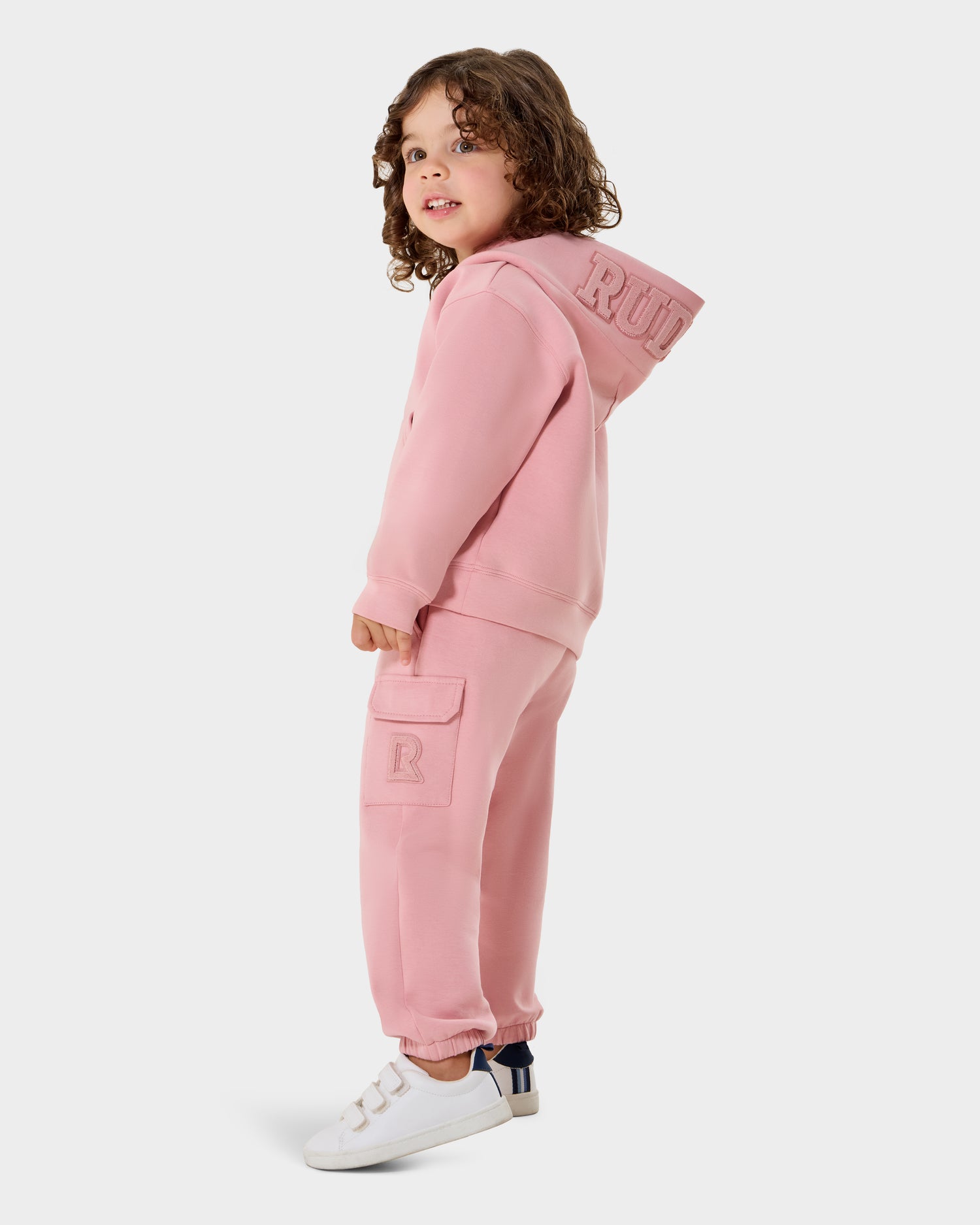 A young child with curly hair wears the RAINEY - 1525002 Light Pink hoodie and matching joggers from Rudsak, paired with white sneakers, smiling against a light background. A perfect stylish kids outfit by Rudsak.