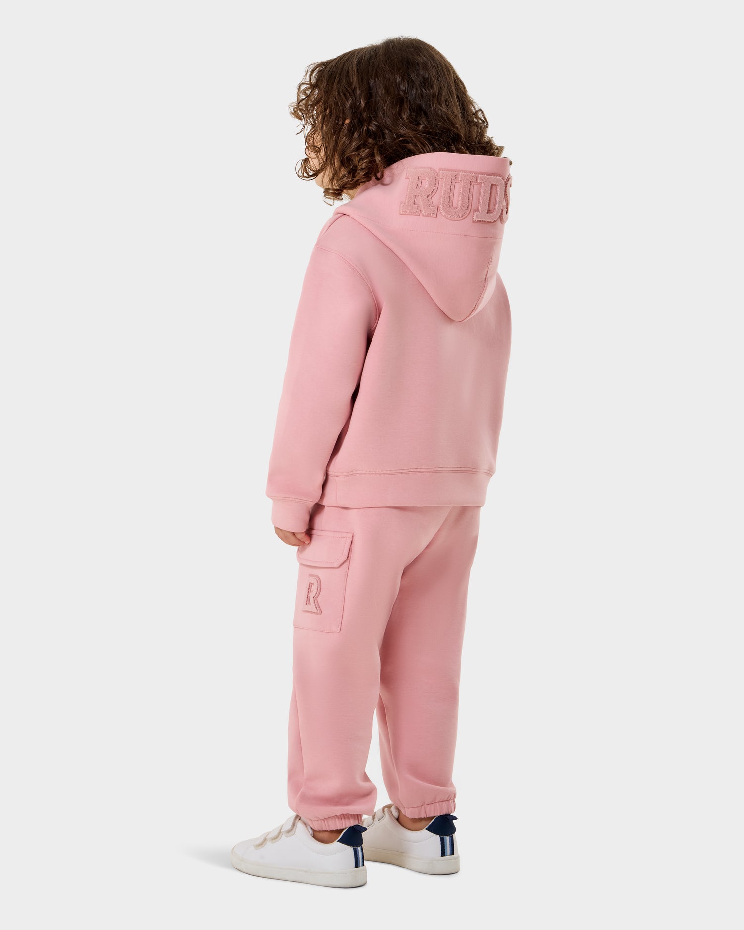 A child with curly hair stands facing away, wearing the Rudsak RAINEY - 1525002 light pink hoodie with "RUDE" on the hood, matching light pink sweatpants with a pocket, and white sneakers with blue heel accents.