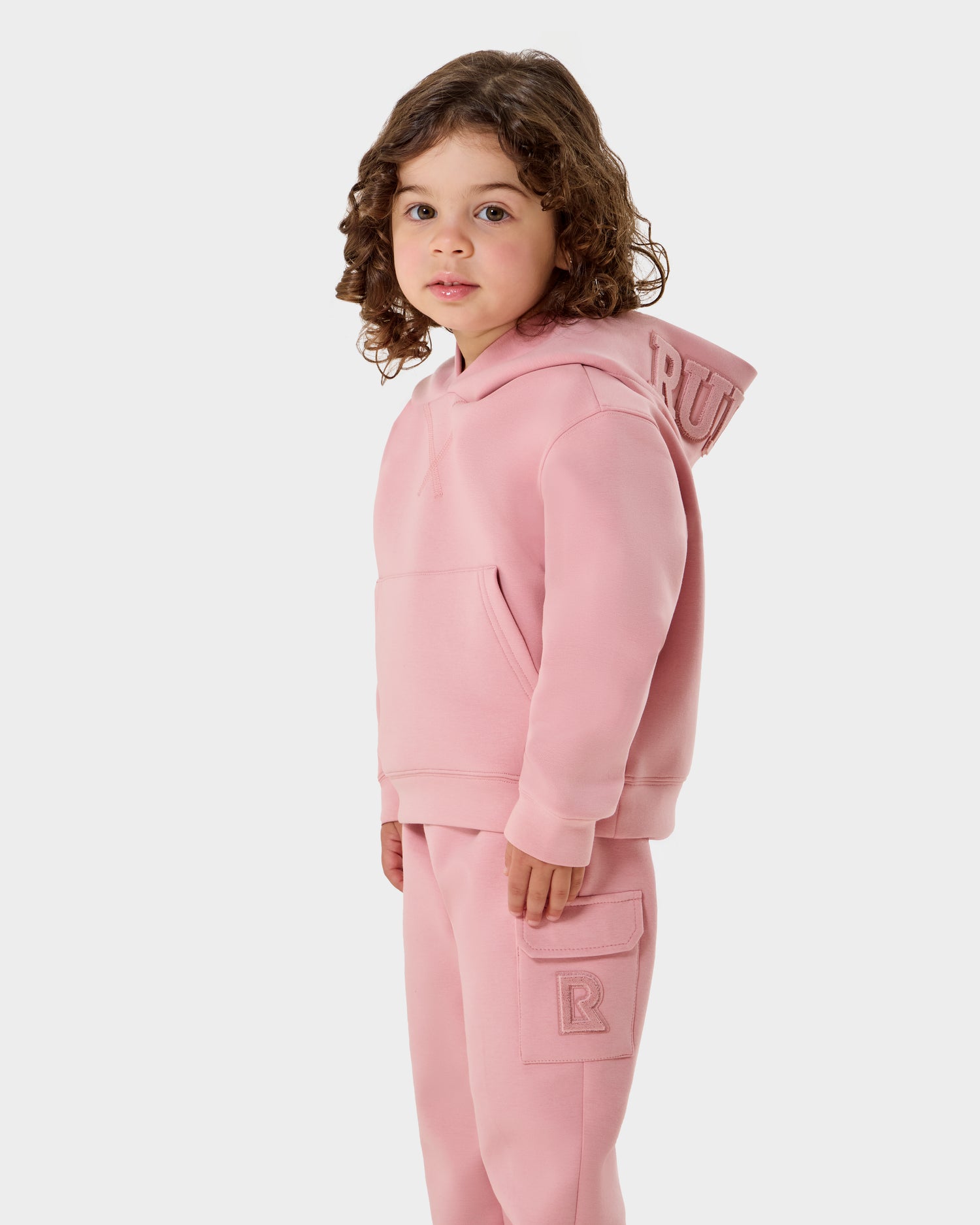 A young child with curly brown hair wears the Rudsak RAINEY - 1525002 Light Pink hoodie and sweatpants, featuring pockets and R details, standing and looking slightly to the side against a plain white background.
