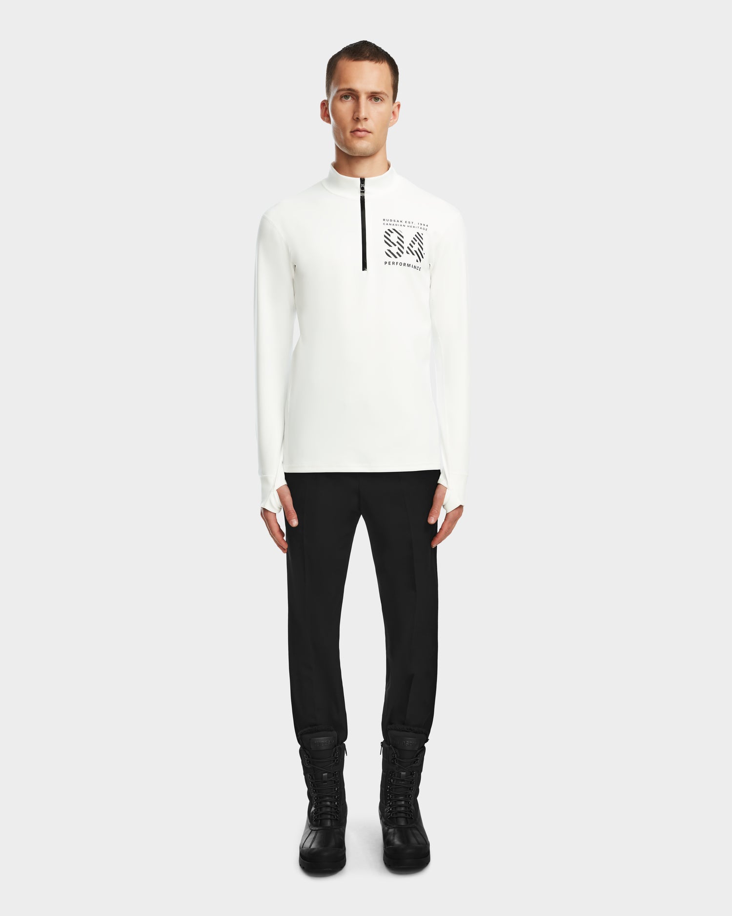 A man stands facing forward wearing the Rudsak RALI - 6524522 Optic White long-sleeve zip-up shirt with black text on the chest, paired with black pants and boots, against a plain light background.