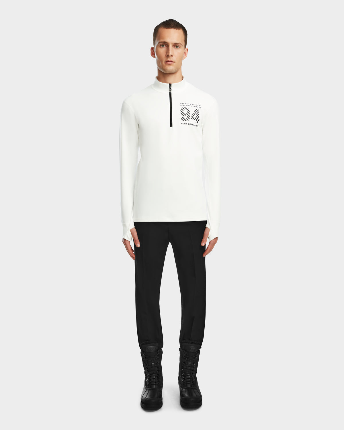A man stands facing forward wearing the Rudsak RALI - 6524522 Optic White long-sleeve zip-up shirt with black text on the chest, paired with black pants and boots, against a plain light background.