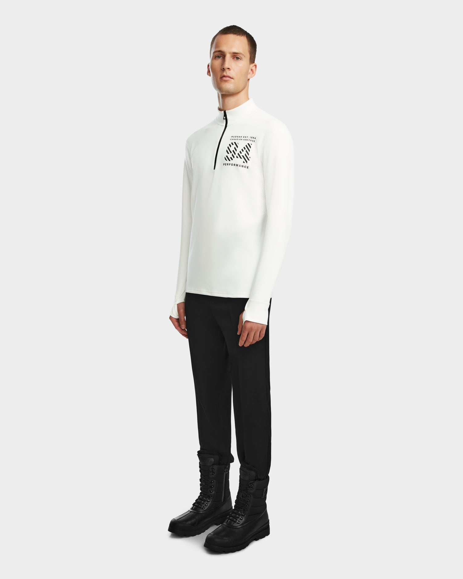A person faces slightly left, wearing the RALI - 6524522 Optic White men’s active top by Rudsak with a zip-neck and black chest graphic, paired with black pants and boots—ideal as a layering shirt for winter sports—against a plain light background.