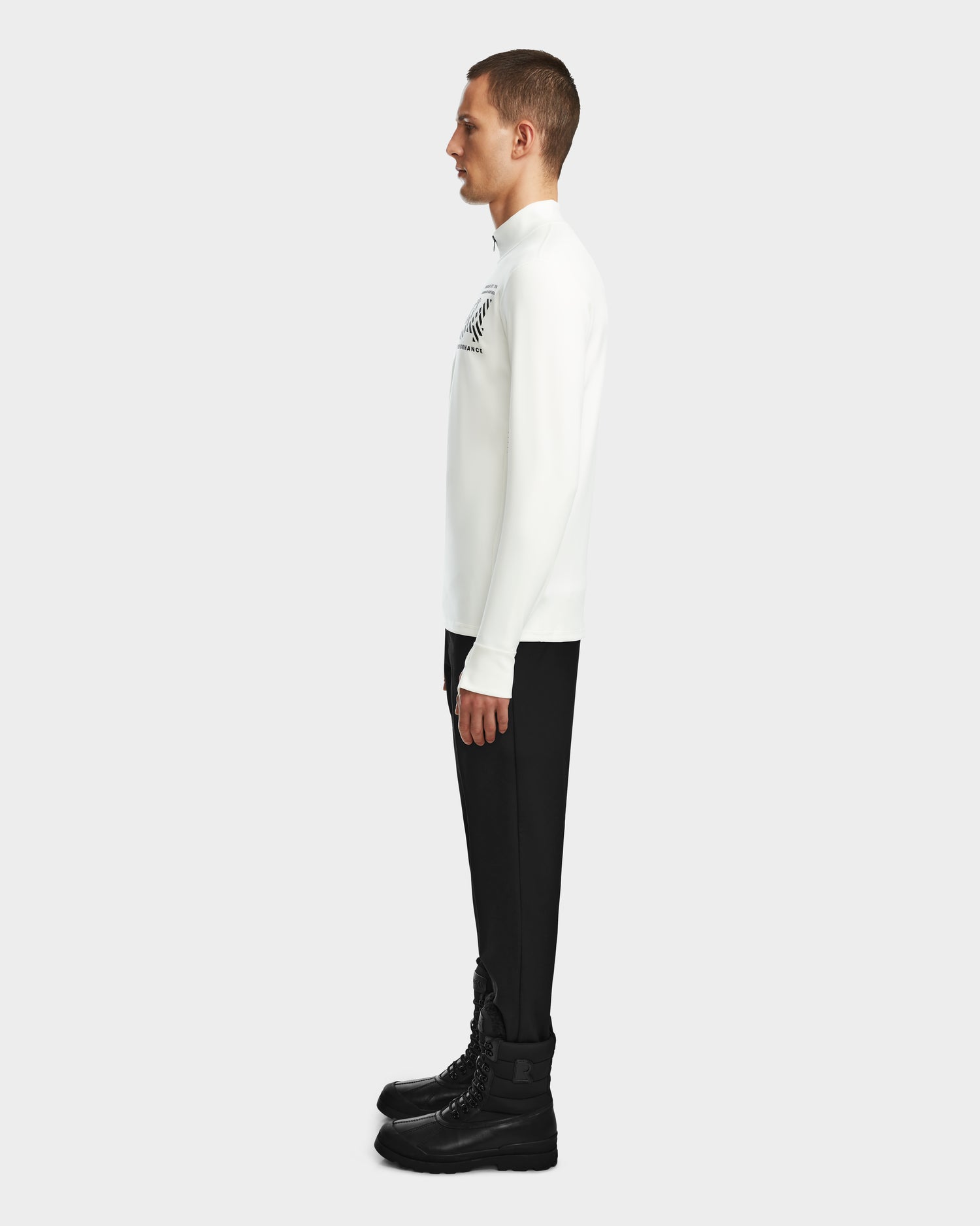 A man stands in profile facing left, wearing the Rudsak RALI - 6524522 Optic White shirt, black pants, and black lace-up boots against a plain light gray background.