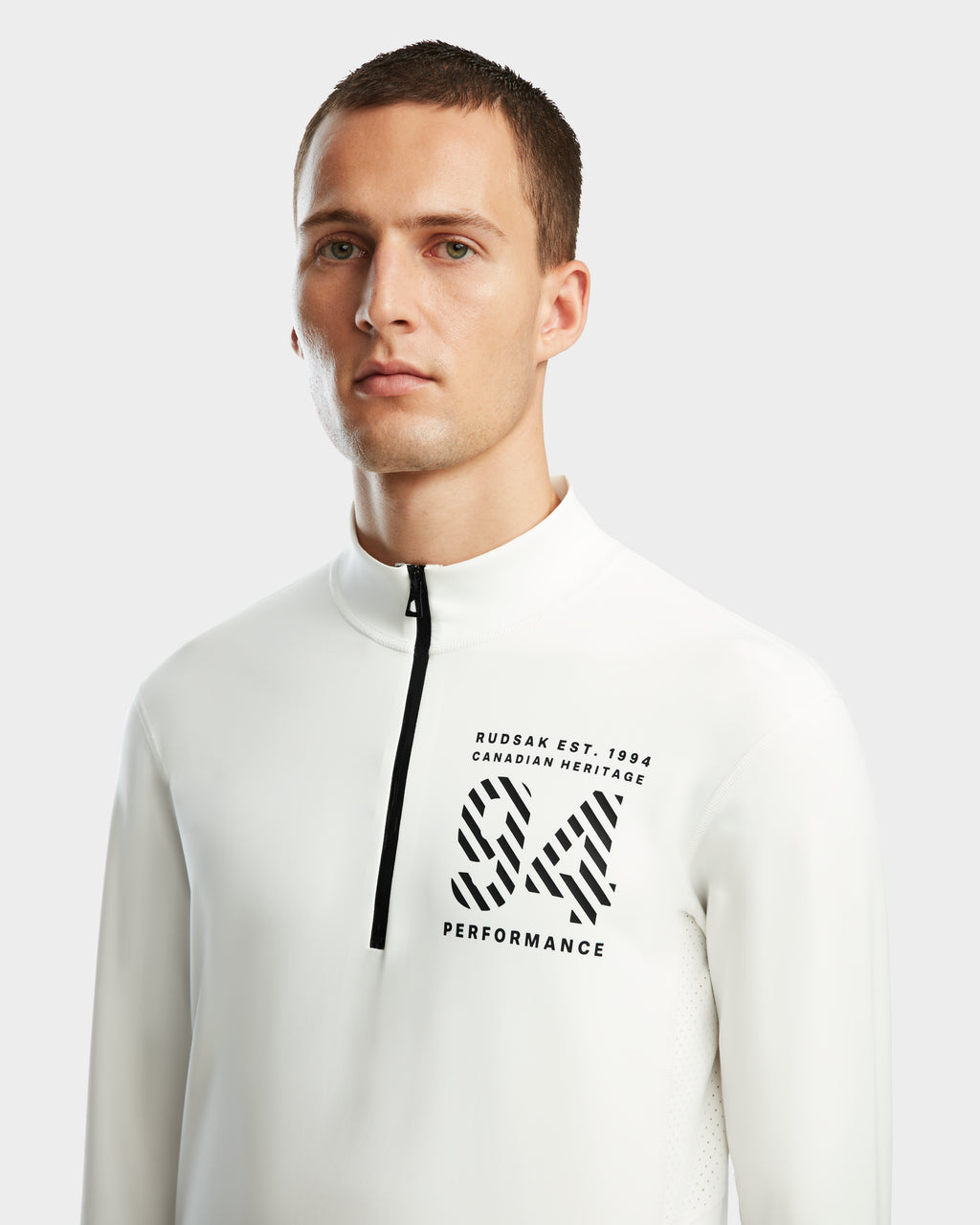 A young man with short brown hair wears Rudsak’s RALI - 6524522 Optic White zip-neck active top, featuring black “RUDSAK EST. 1994 CANADIAN HERITAGE” text and a bold SX PERFORMANCE logo, ideal for layering or winter sports.