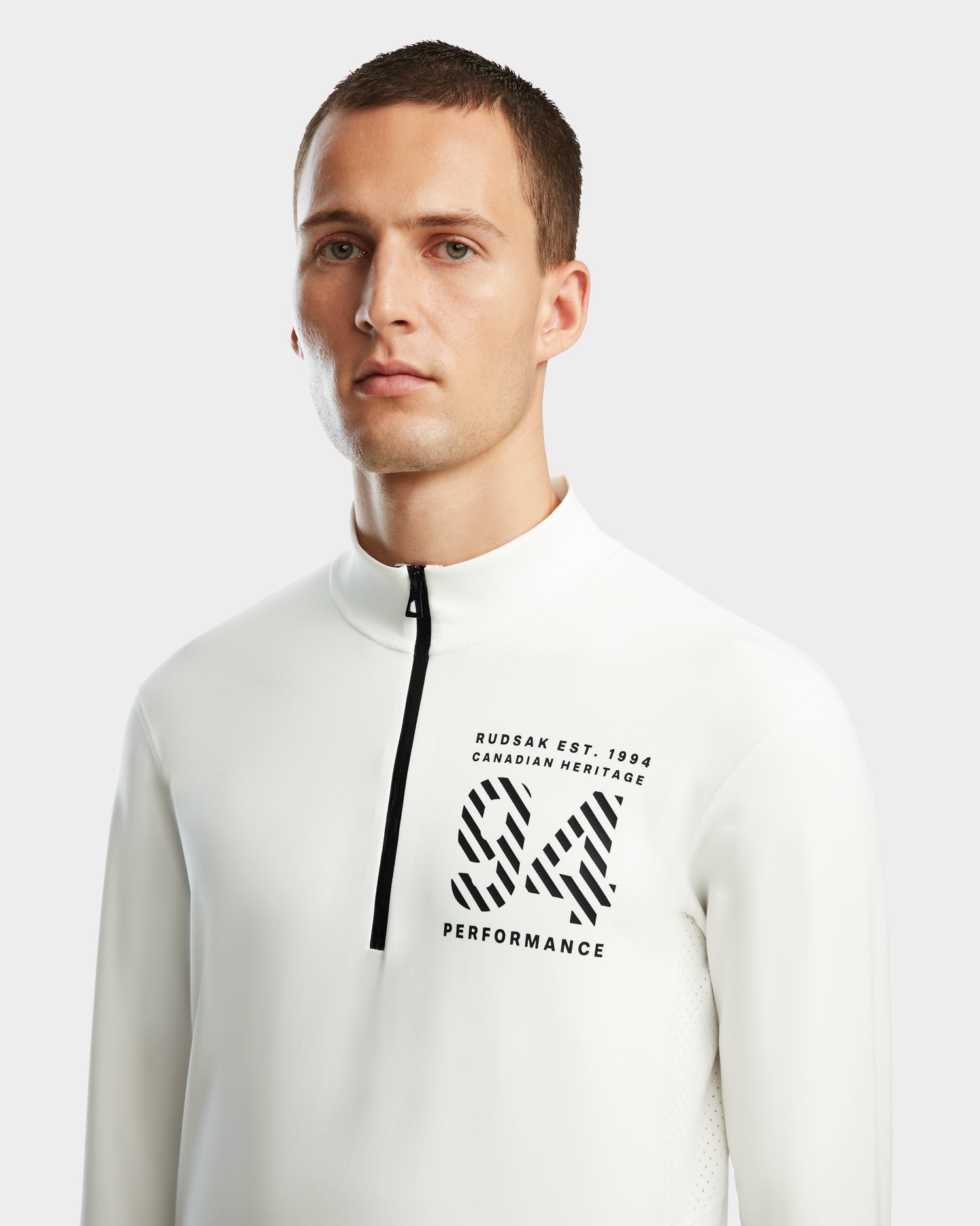 A young man with short brown hair wears Rudsak’s RALI - 6524522 Optic White zip-neck active top, featuring black “RUDSAK EST. 1994 CANADIAN HERITAGE” text and a bold SX PERFORMANCE logo, ideal for layering or winter sports.