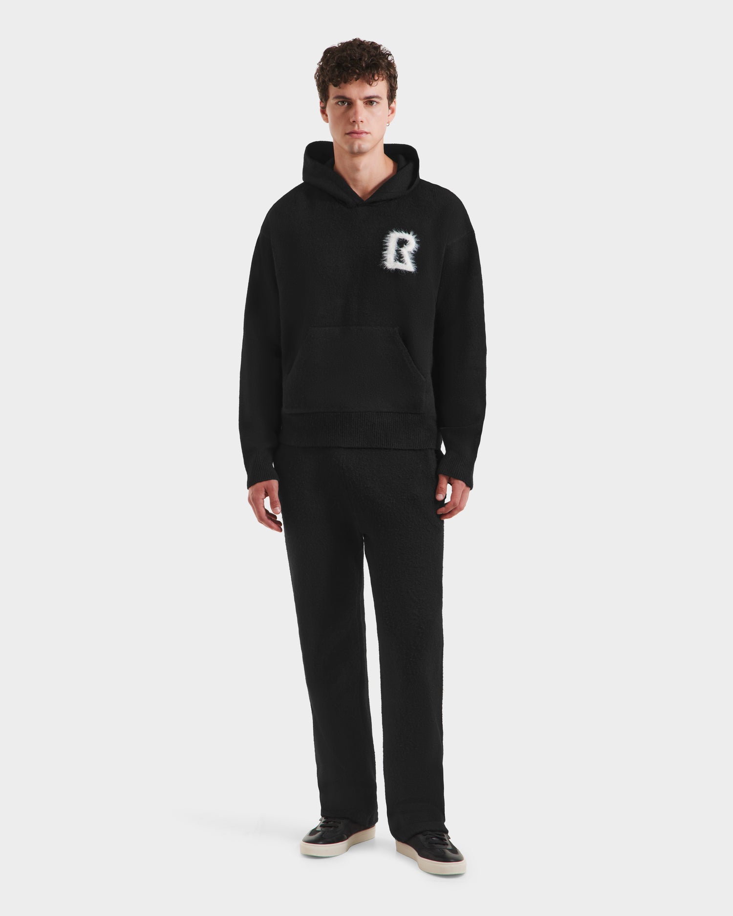 A person wears the Rudsak RAPHAEL - 6525643 Black men’s hoodie with a white R on the chest, paired with matching black pants and black sneakers with white soles, for a relaxed look against a plain white background.