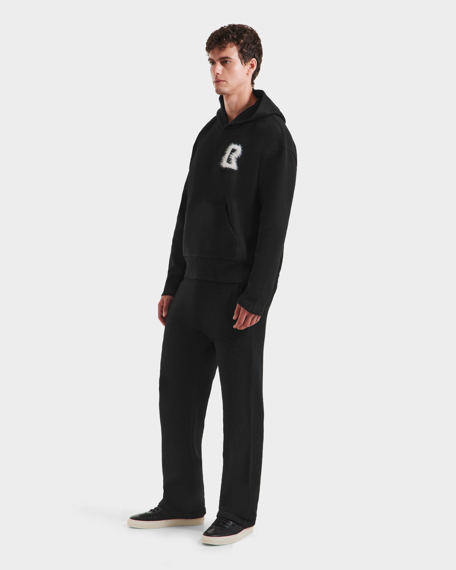 A young man with short curly hair models the RAPHAEL - 6525643 Black men’s hoodie by Rudsak, featuring a large R on the chest and matching black sweatpants, paired with black sneakers against a plain white background.