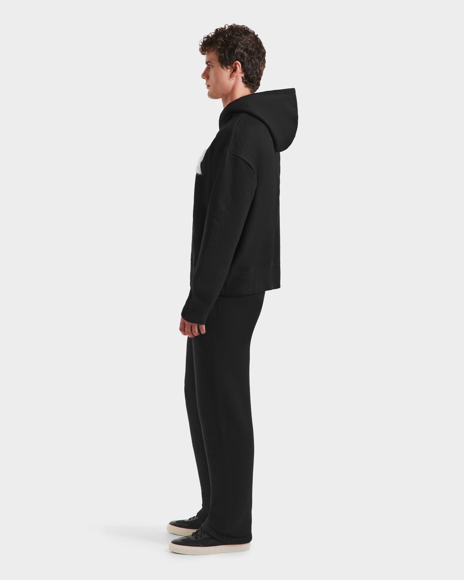 A person with short curly hair stands in profile facing left, wearing the RAPHAEL - 6525643 Black men’s hoodie by Rudsak, black pants, and black sneakers with white soles against a plain white background for a casual streetwear look.