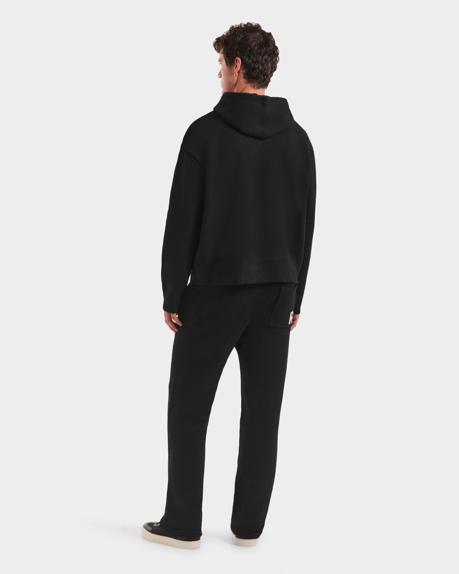 A person with short curly hair faces away, wearing the RAPHAEL - 6525643 Black men’s hoodie and pants by Rudsak as loungewear with light sneakers, against a plain white background.