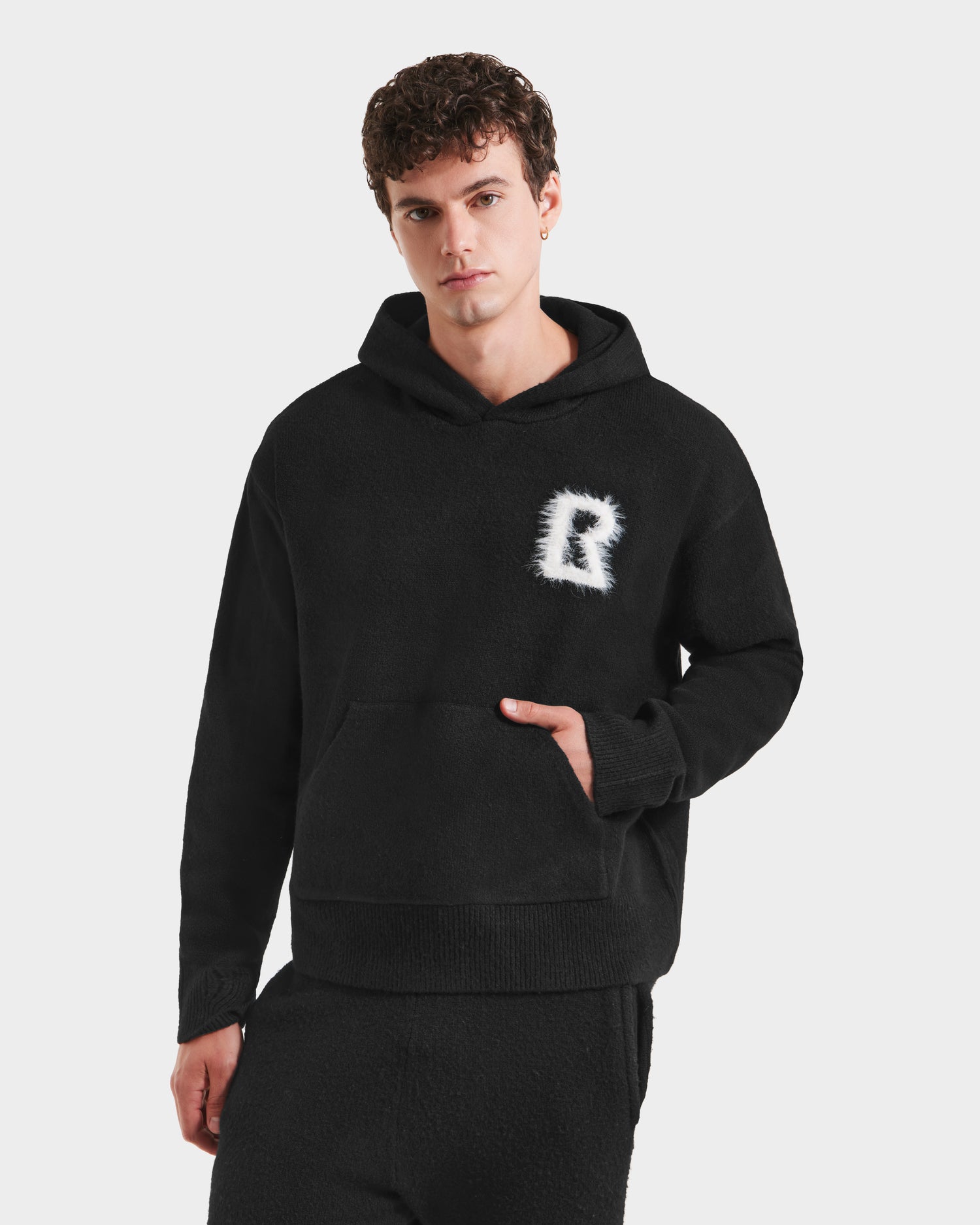 A young man models the Rudsak RAPHAEL - 6525643 Black men’s hoodie with a fuzzy white R logo and matching black loungewear pants, standing against a plain white background with one hand in his pocket.