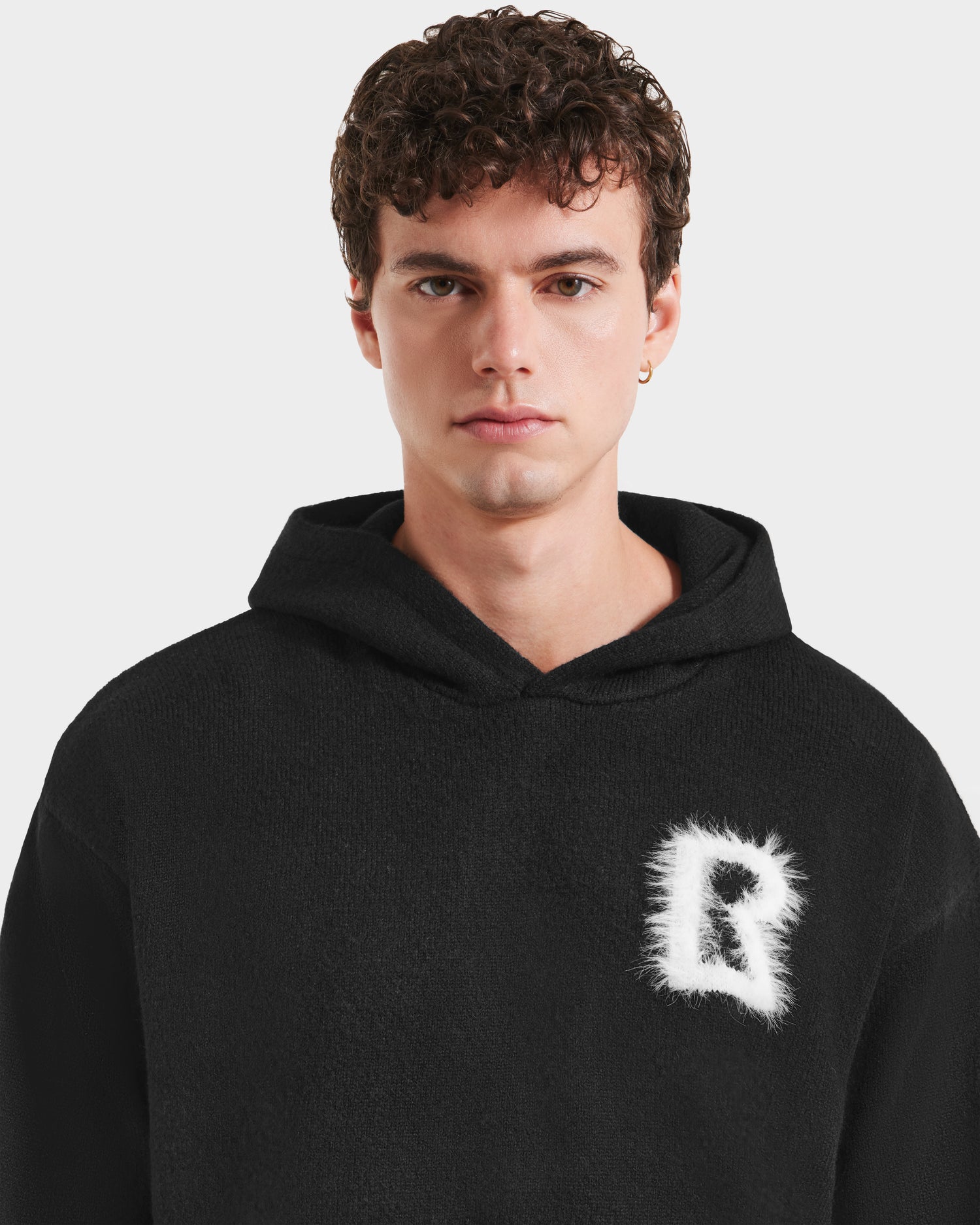 A young man with short curly hair wears the RAPHAEL - 6525643 Black hoodie by Rudsak, featuring a white fuzzy letter R on the left chest. He faces forward with a neutral expression against a plain light background.