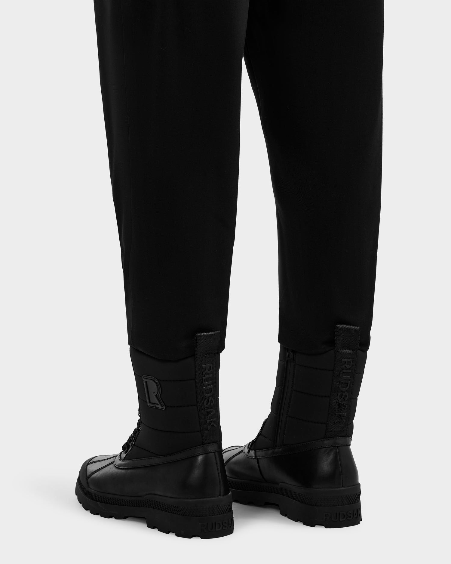 A person wears black pants and REAGAN - 6224501 Black waterproof lace-up leather boots from Rudsak, ideal for winter. The boots feature embossed Rudsak branding on heel tabs and sides. Photo shows only lower legs and feet from behind.
