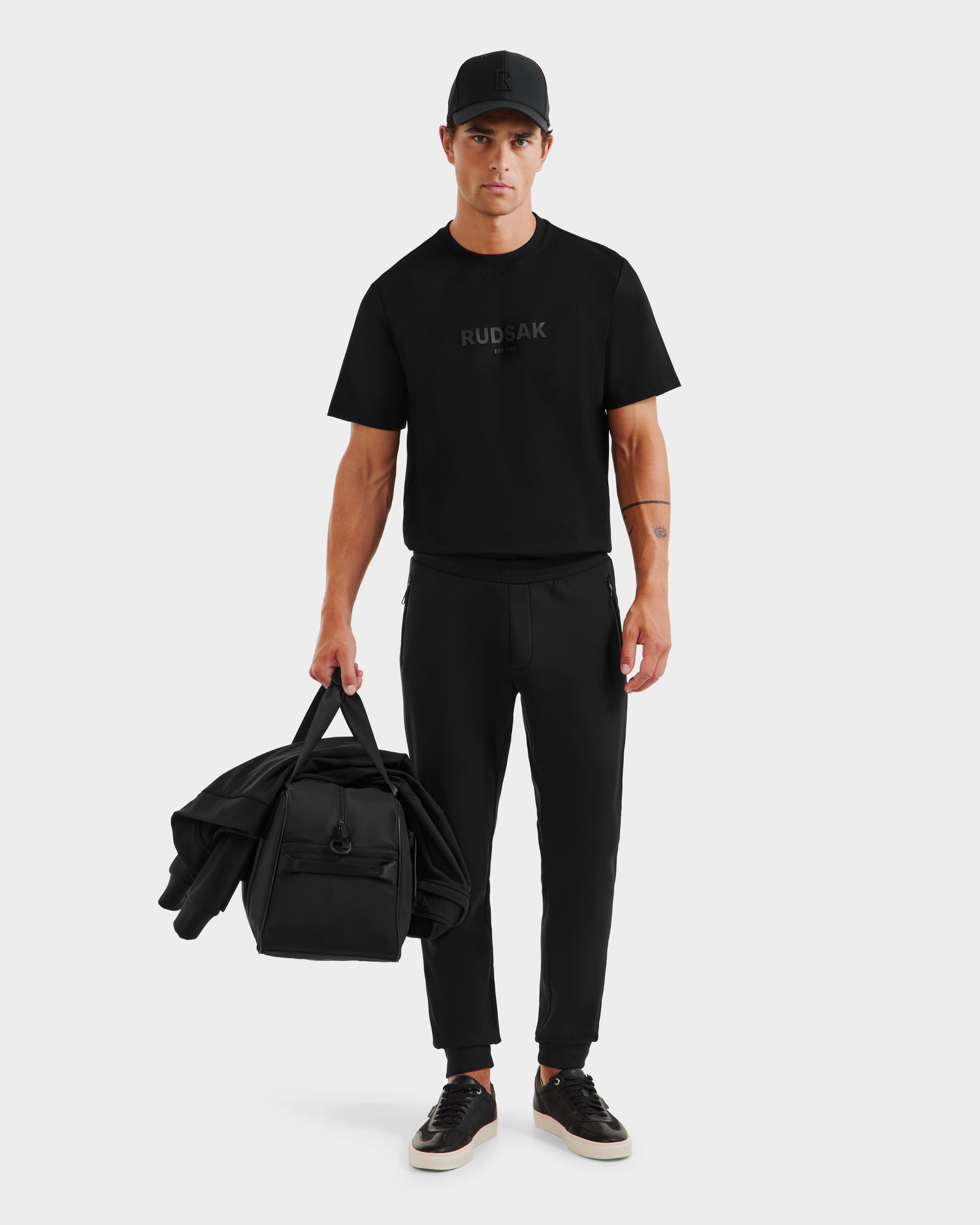 A man wearing the Rudsak REGIS - 6525582 Black outfit stands against a plain white background, holding a matching black jacket and duffel bag in his left hand.