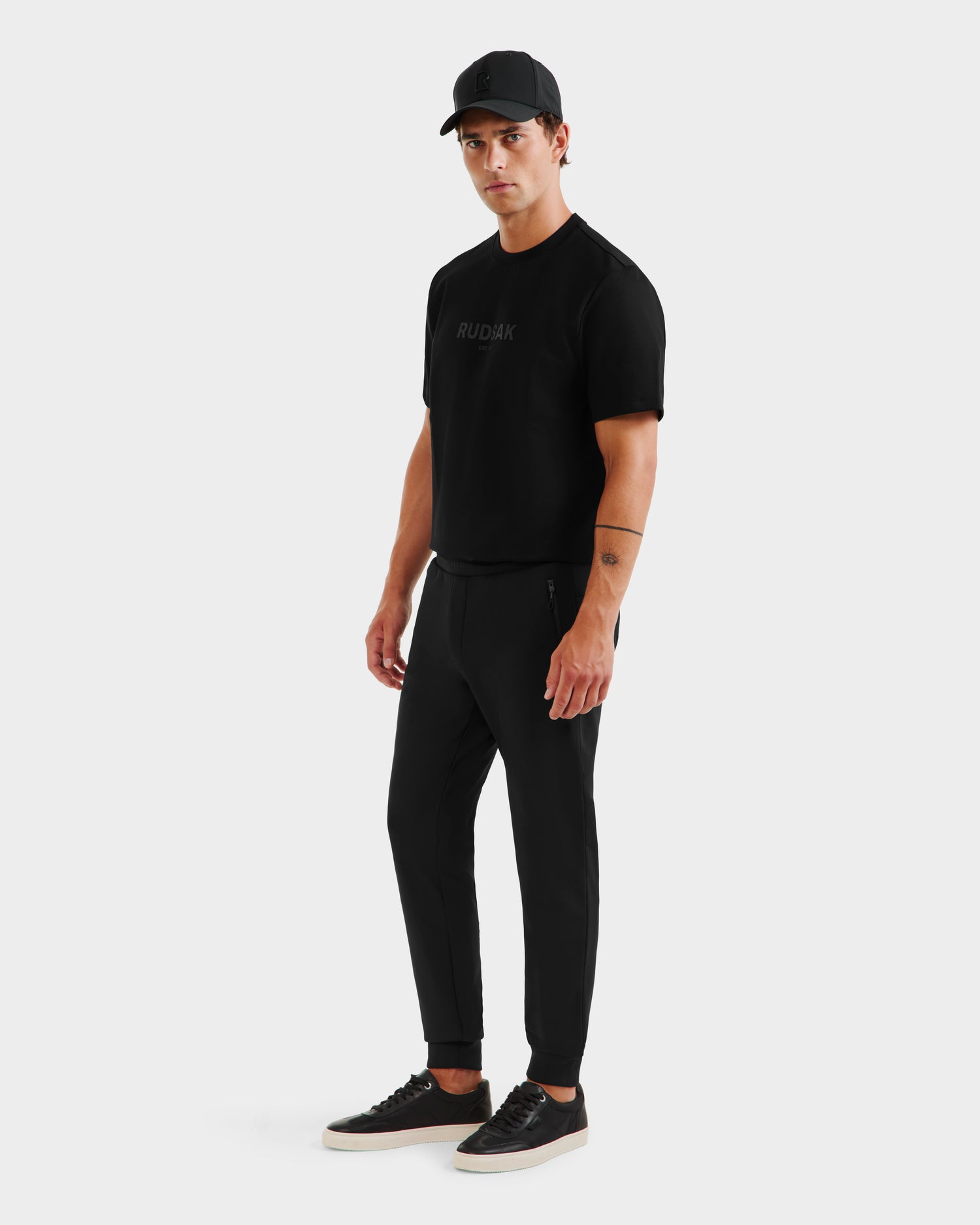 A man stands against a plain background wearing a black cap, black t-shirt, and REGIS - 6525582 Black jogger pants by Rudsak with sneakers. He faces slightly to the side, arms relaxed—a sleek look styled with athletic joggers.