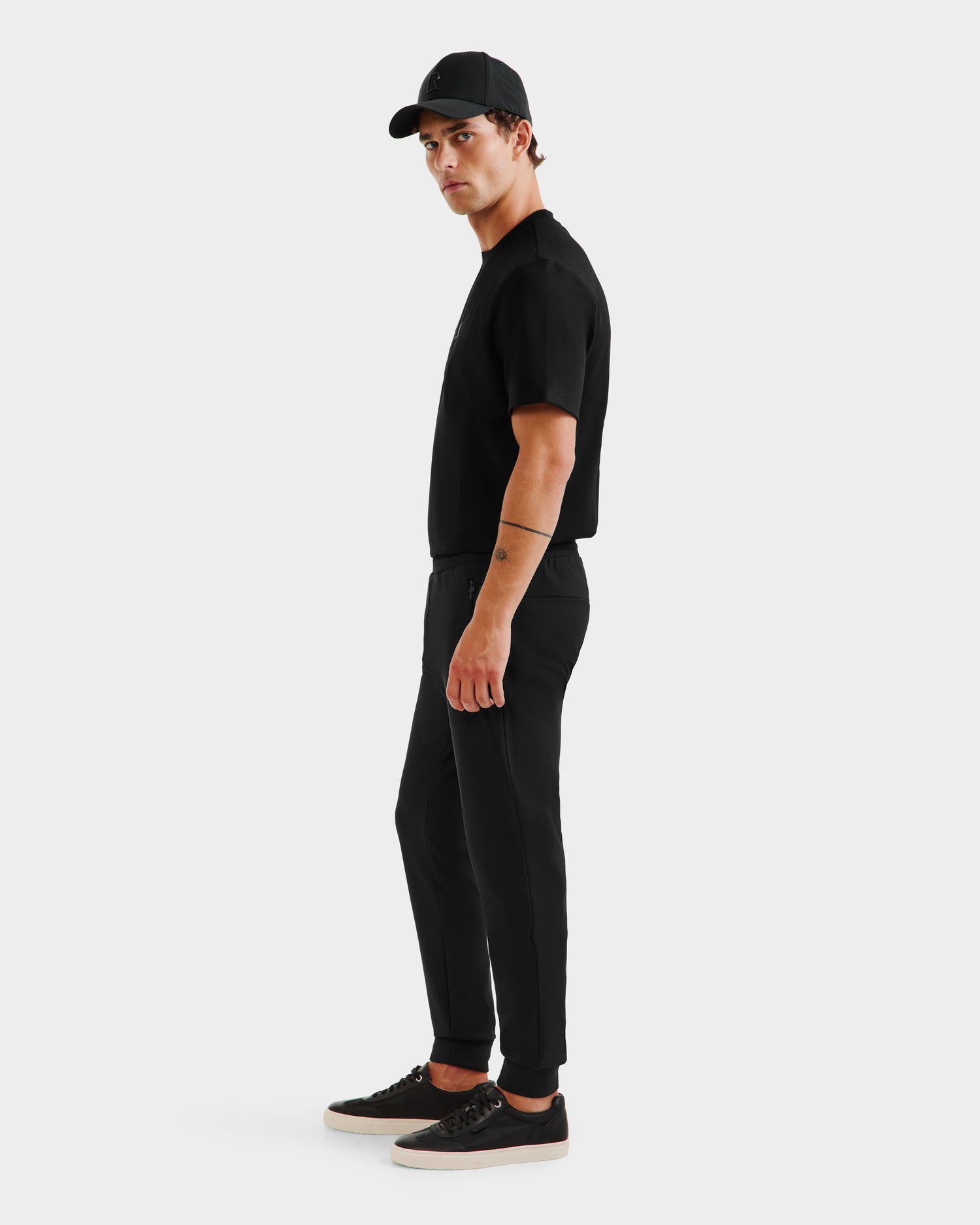 A man wearing Rudsak’s REGIS - 6525582 Black jogger pants, a black cap, and a black t-shirt stands sideways, looking at the camera against a plain white background.