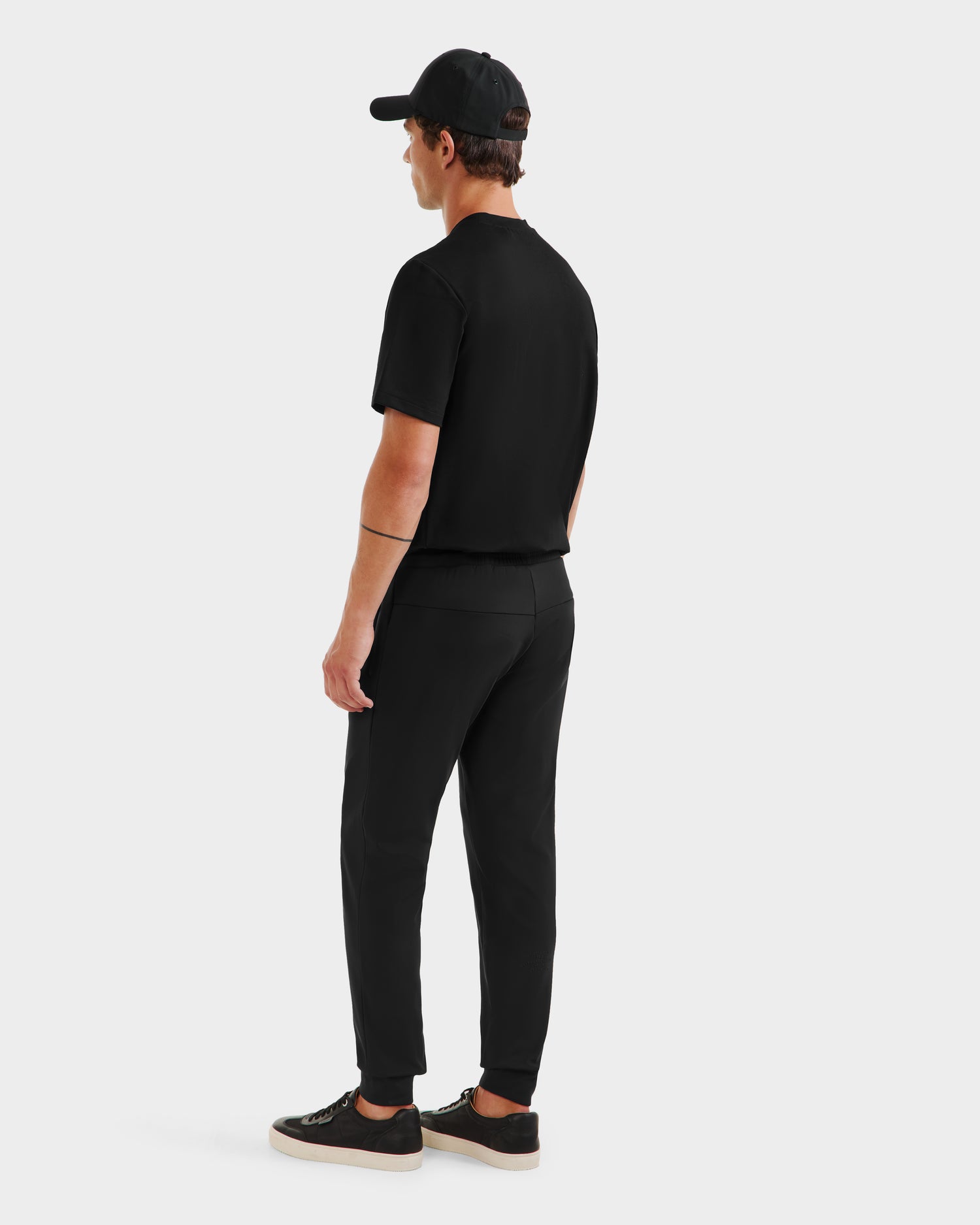 A man, seen from behind, wears the Rudsak REGIS - 6525582 Black baseball cap, black short-sleeve shirt, athletic joggers, and black sneakers with white soles against a plain white background.
