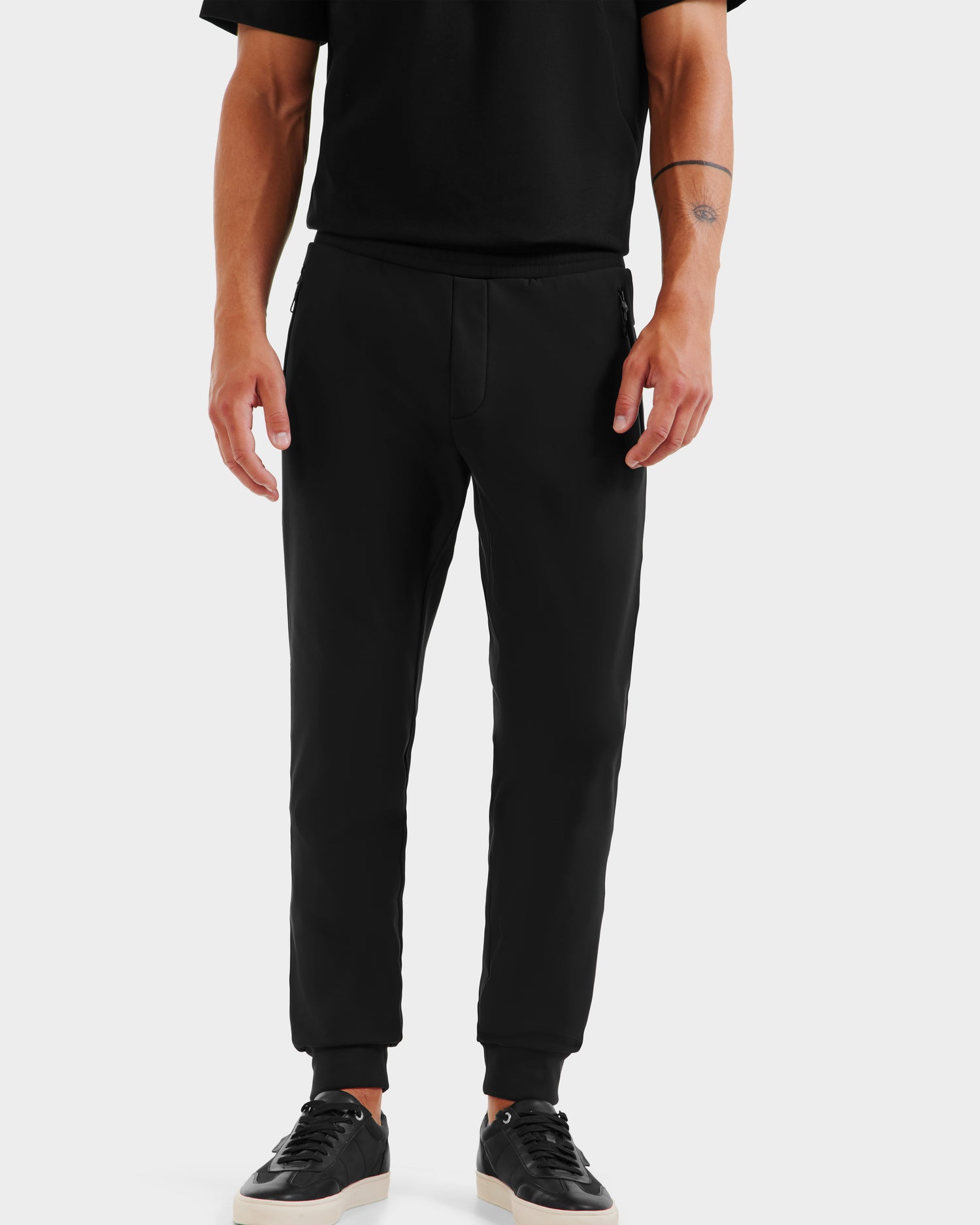 A person stands against a plain white background, wearing Rudsak REGIS - 6525582 Black men’s performance jogger pants, a black T-shirt, and black sneakers with white soles. The person’s face is not visible.
